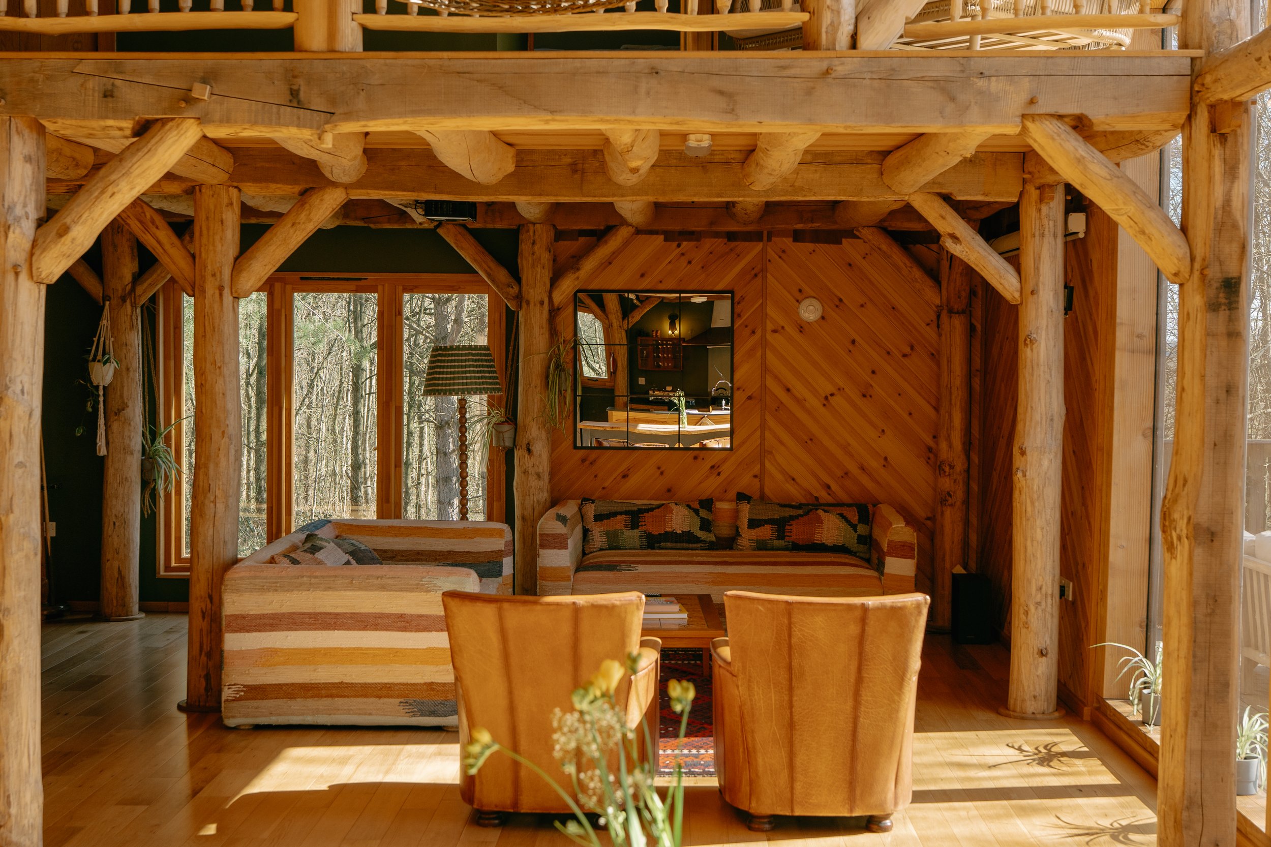 Cosy living room with wooden logs and rustic decor, featuring striped sofas, leather chairs, and a large window view of the forest.