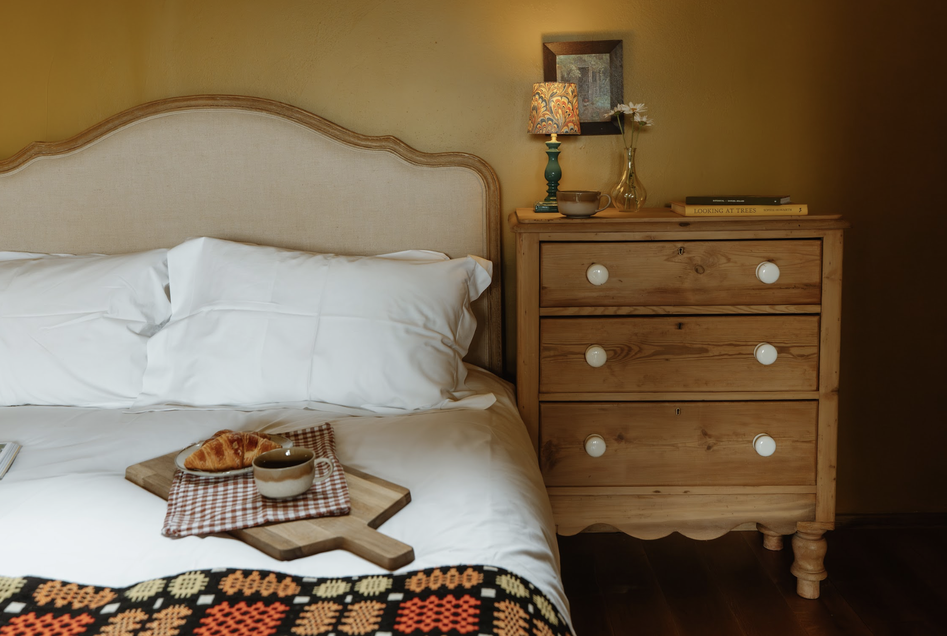 A cozy bedroom with a bed, white bedding, and a tray with a croissant and a cup on it. Next to the bed is a wooden dresser with a lamp, a vase with flowers, and books on top. The wall behind has a framed picture.
