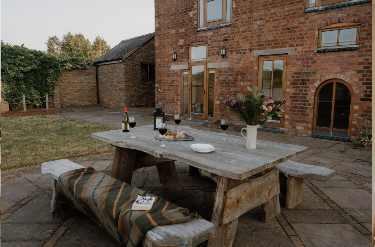 Outdoor dining area with wooden table set with glasses of red wine, a bottle of wine, a cheese platter, a lantern, and a flower vase with pink and white flowers in front of a brick house with wooden framed windows and doors.