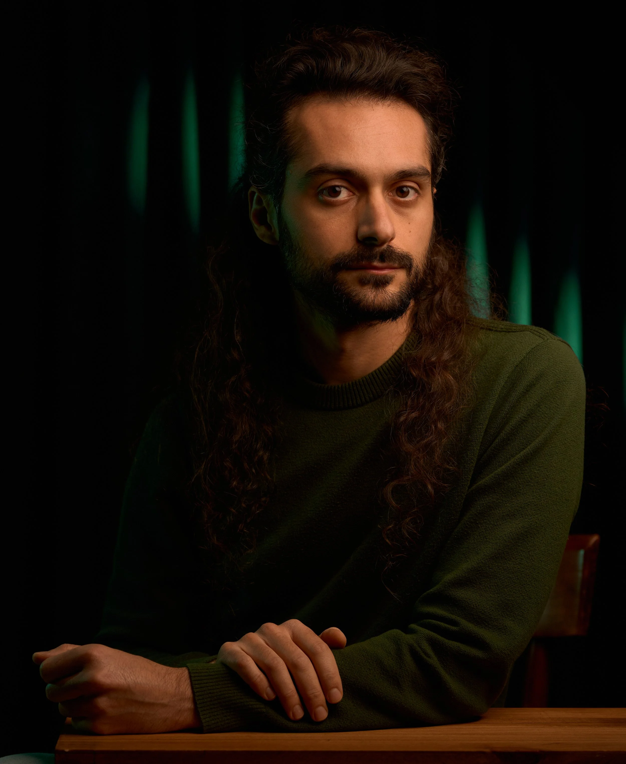 Ritratto di Manuel Montesano, fotografo, videografo, editor video, colorist. Uomo con capelli lunghi e ricci, barba, che indossa una maglia verde scuro, seduto di fronte a un tavolo di legno, con sfondo scuro con luci verdi