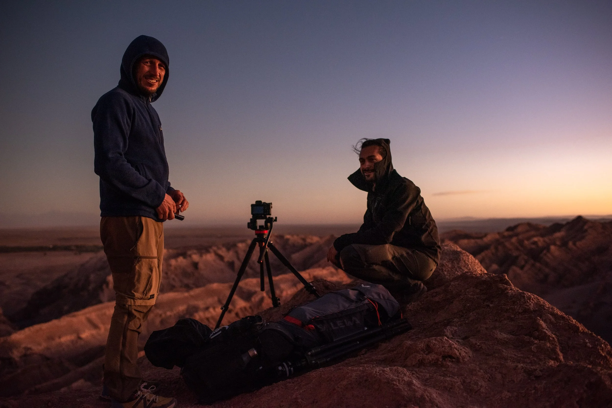 Manuel Montesano e Davide Monteleone, nel deserto di Atacama, Cile, con panorama di canyon dietro di loro. Uno di loro è in piedi, l'altro è seduto su una roccia, ci sono tre treppiedi e attrezzature fotografiche, e uno zaino sul terreno.