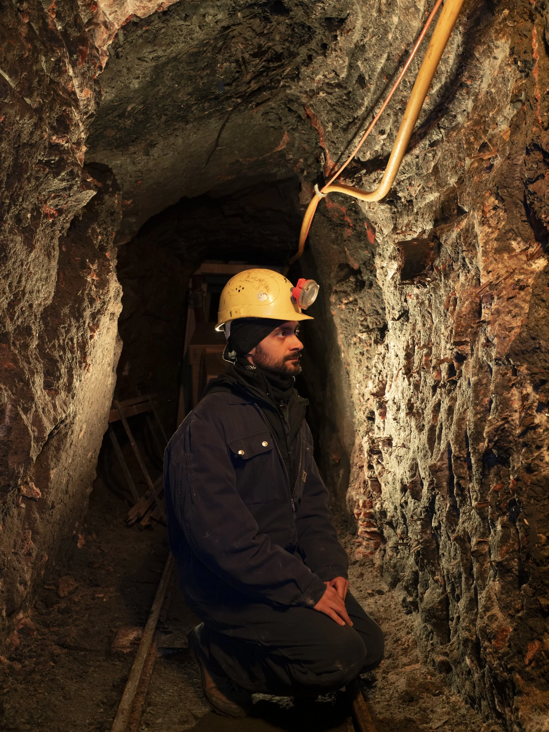 Uomo che lavora in una miniera o tunnel, indossa casco giallo con torcia, abbigliamento da lavoro nero, seduto o in posizione di preghiera sulle rotaie.