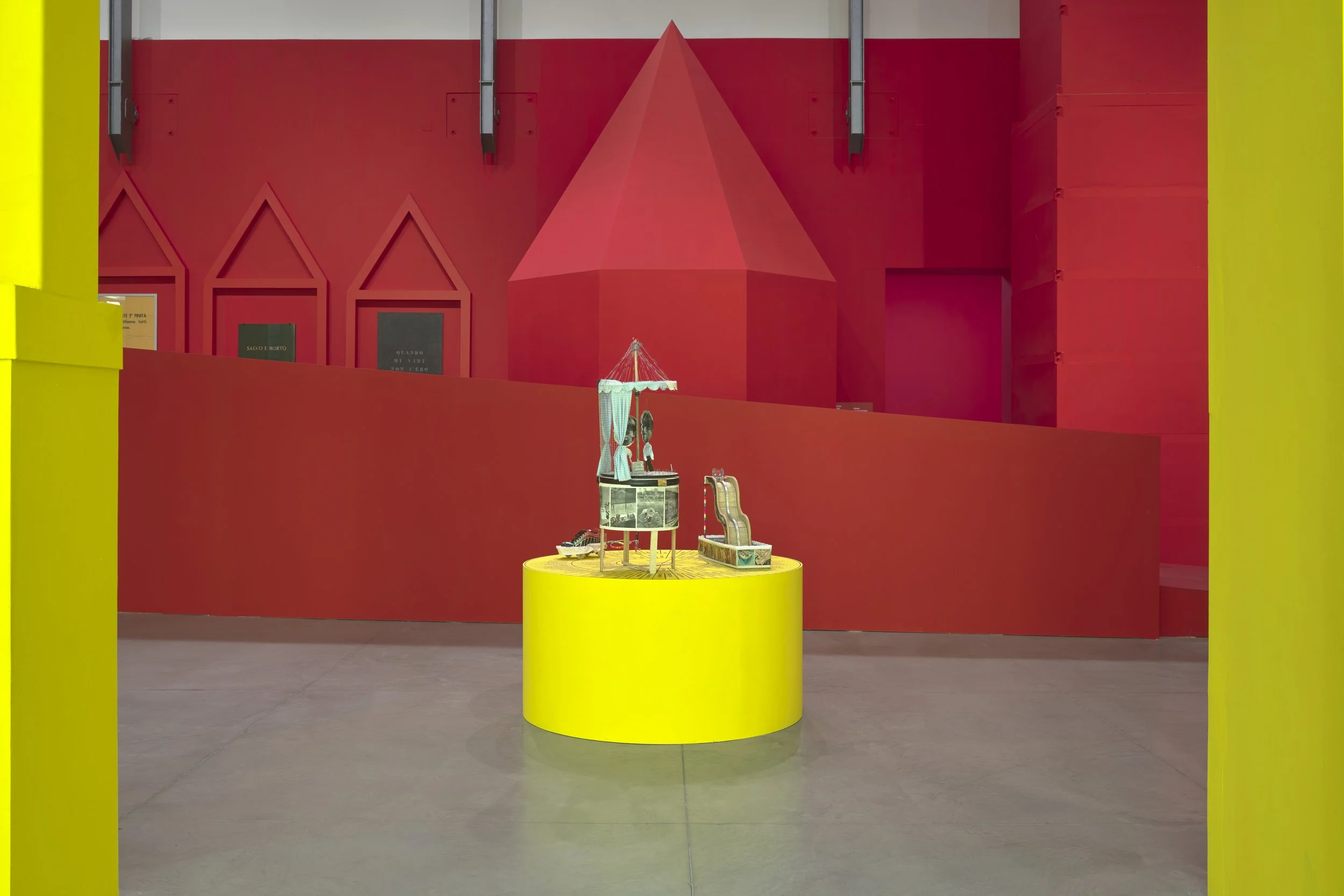 Oggetto di esposizione in una mostra con sfondo rosso e pareti gialle, su un piedistallo giallo, che sembra una collezione di piccoli oggetti, tra cui un'ombrellone, giocattoli e altri piccoli pezzi.