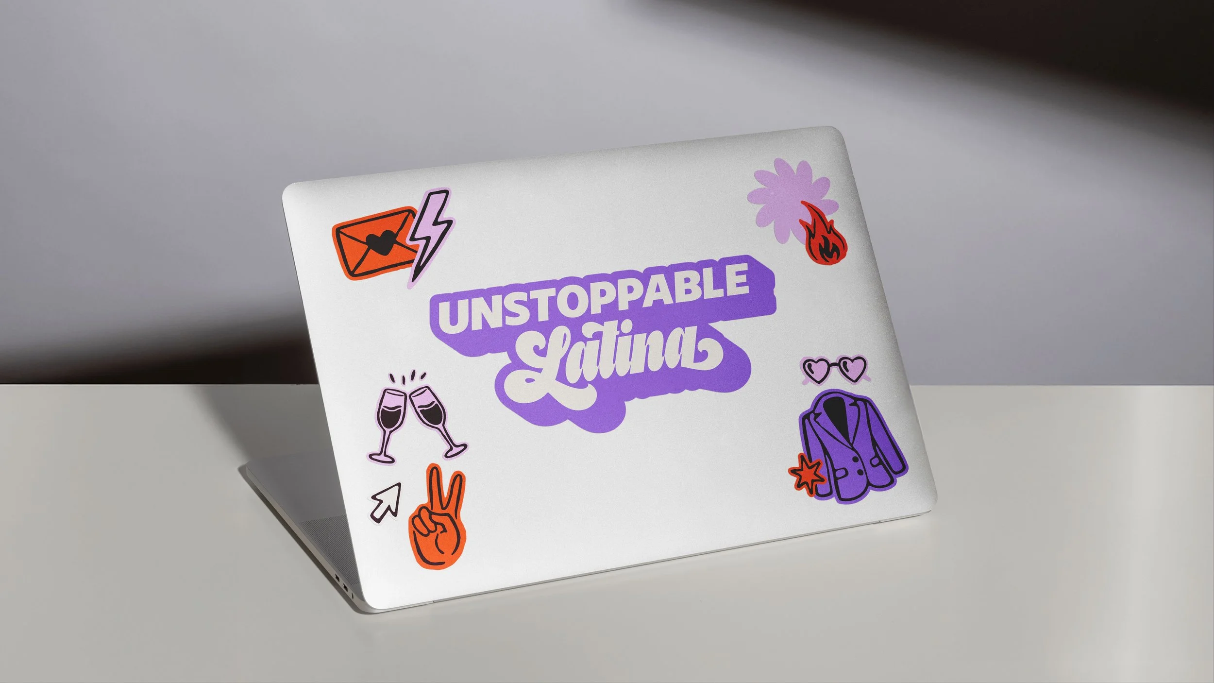 Grey laptop viewed from the back at an angle on a white table against a grey background, decorated with on-brand stickers in violet, plum, orange, pink, and off-white, including a large Unstoppable Latina logo sticker centered on the laptop’s back.
