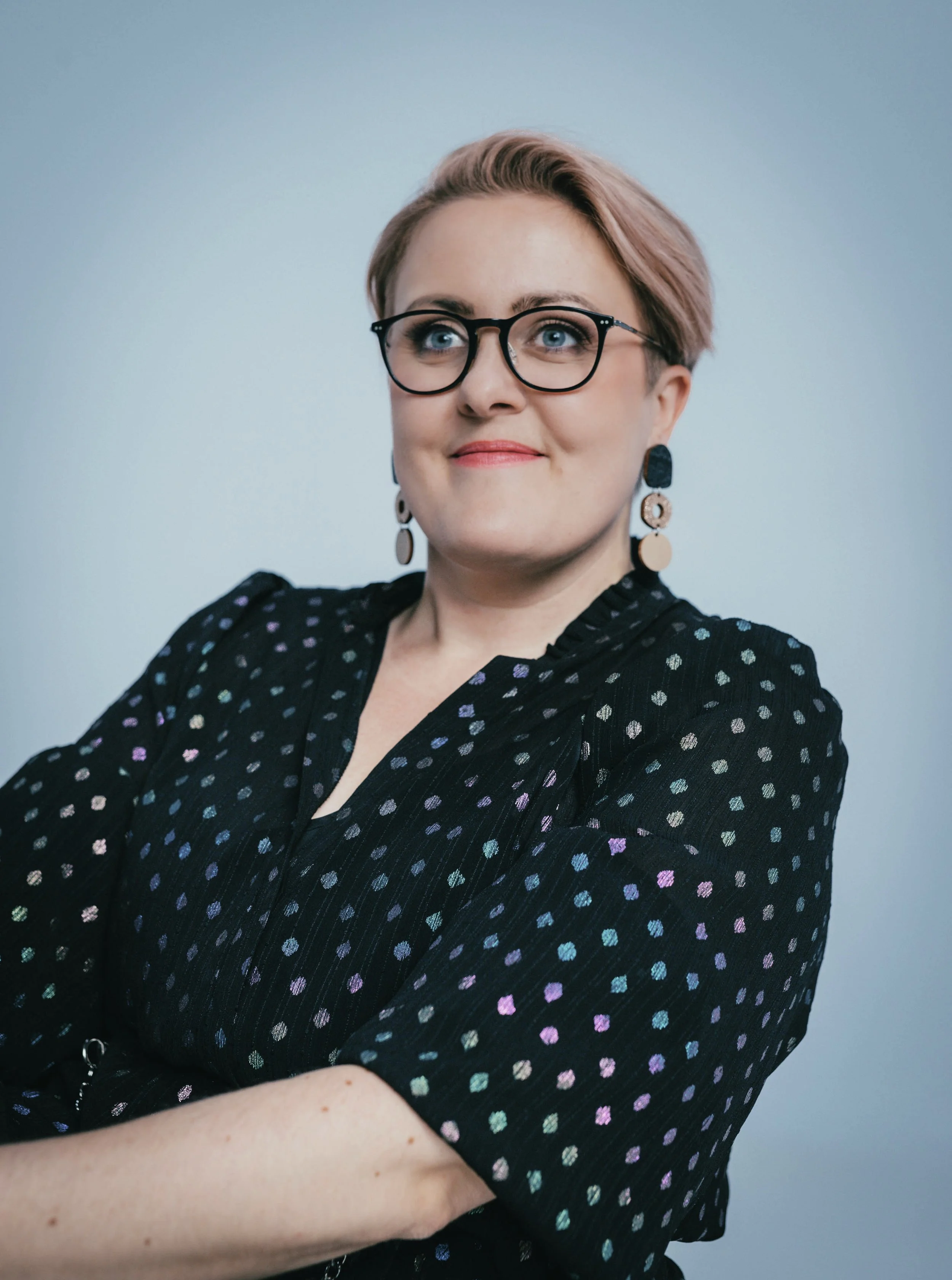 A woman with short, light brown hair styled to the side, wearing black-rimmed glasses, colorful polka dot dress, and multicolored dangling earrings. She is standing against a plain background, with her arms crossed and a confident expression on her face.