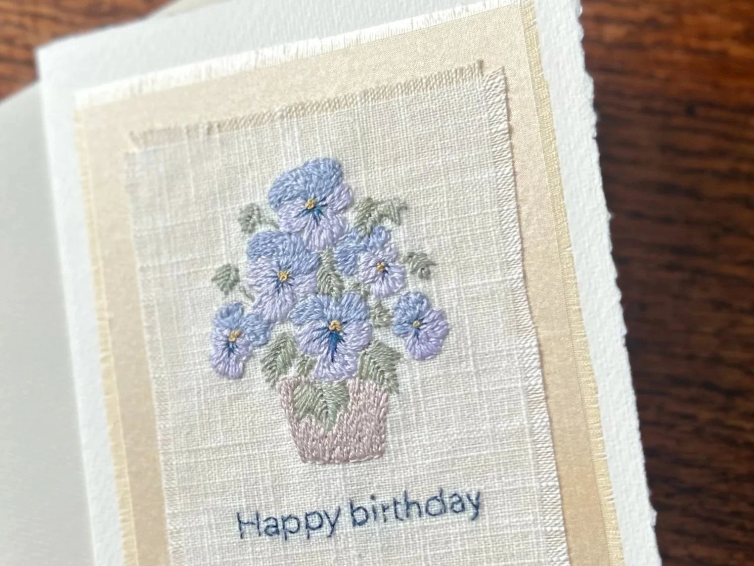 Embroidery of a vase with pansies and green leaves, with the words "Happy birthday" embroidered below.