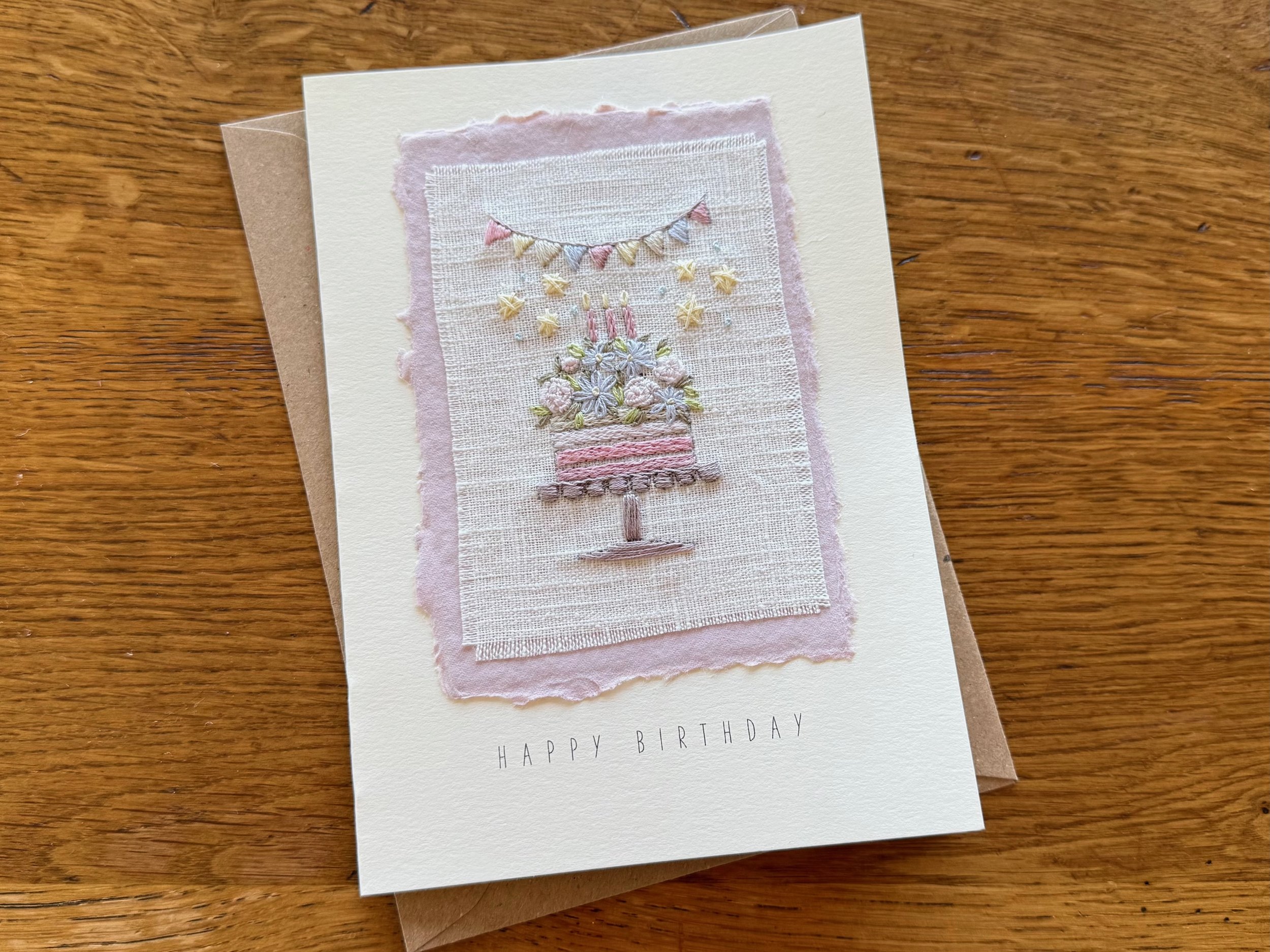 A birthday greeting card with embroidered design of a cake with flowers, candles, and bunting on fabric, on a wooden surface.