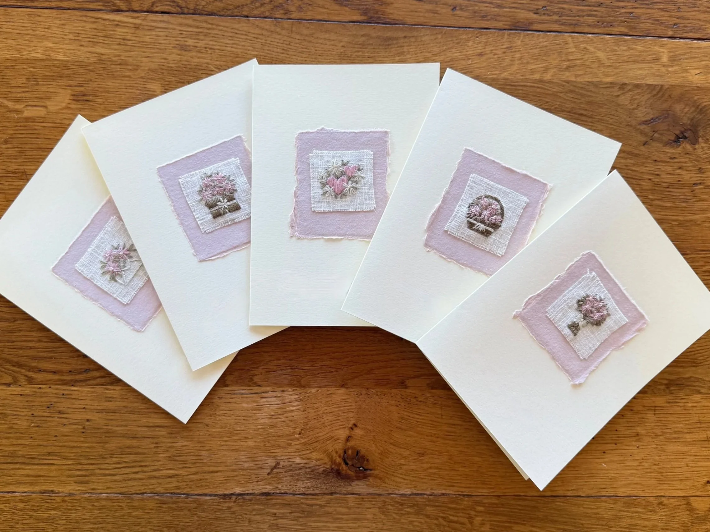Five greeting cards with embroidered floral designs mounted on pink handmade paper and placed on a cream greeting card.