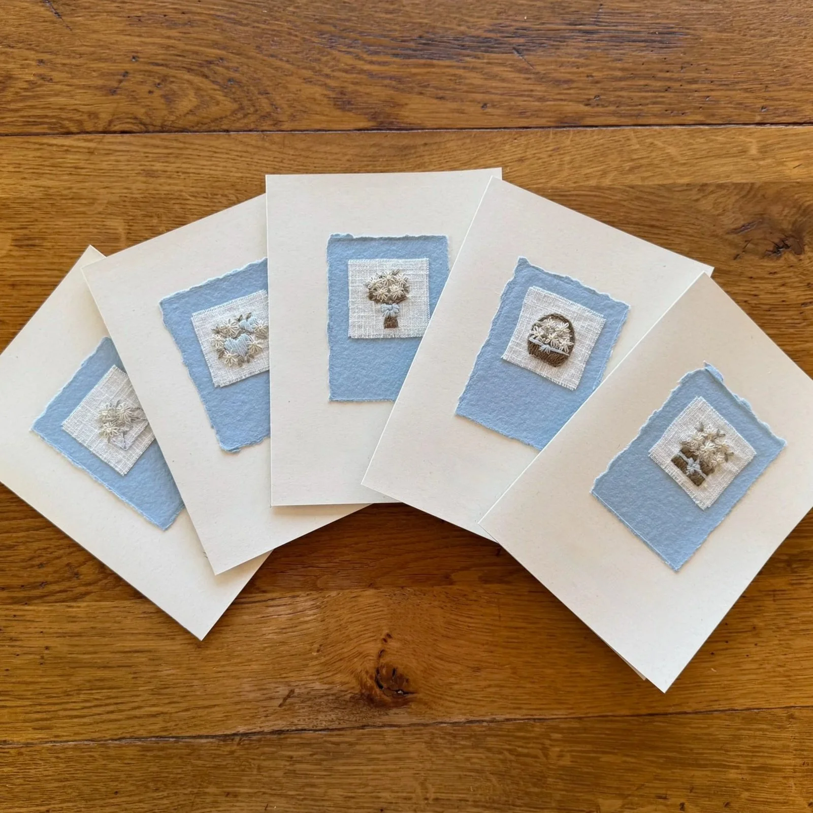 Five greeting cards with embroidered floral designs mounted on blue handmade paper and placed on a cream greeting card.