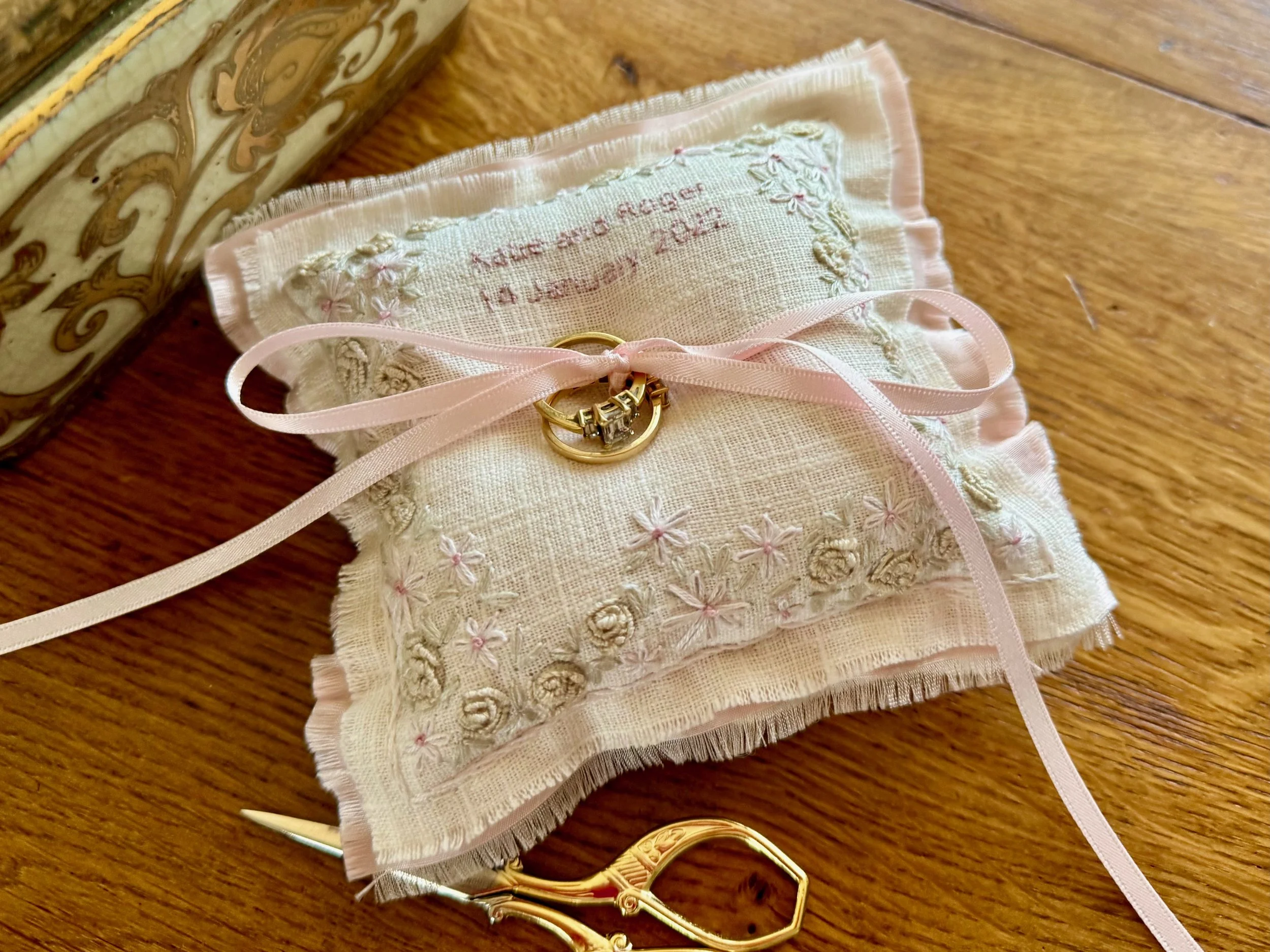A hand embroidered wedding ring pillow with flower design a pink ribbon to tie the wedding rings; personalised with the wedding date and the names of the bride and groom.
