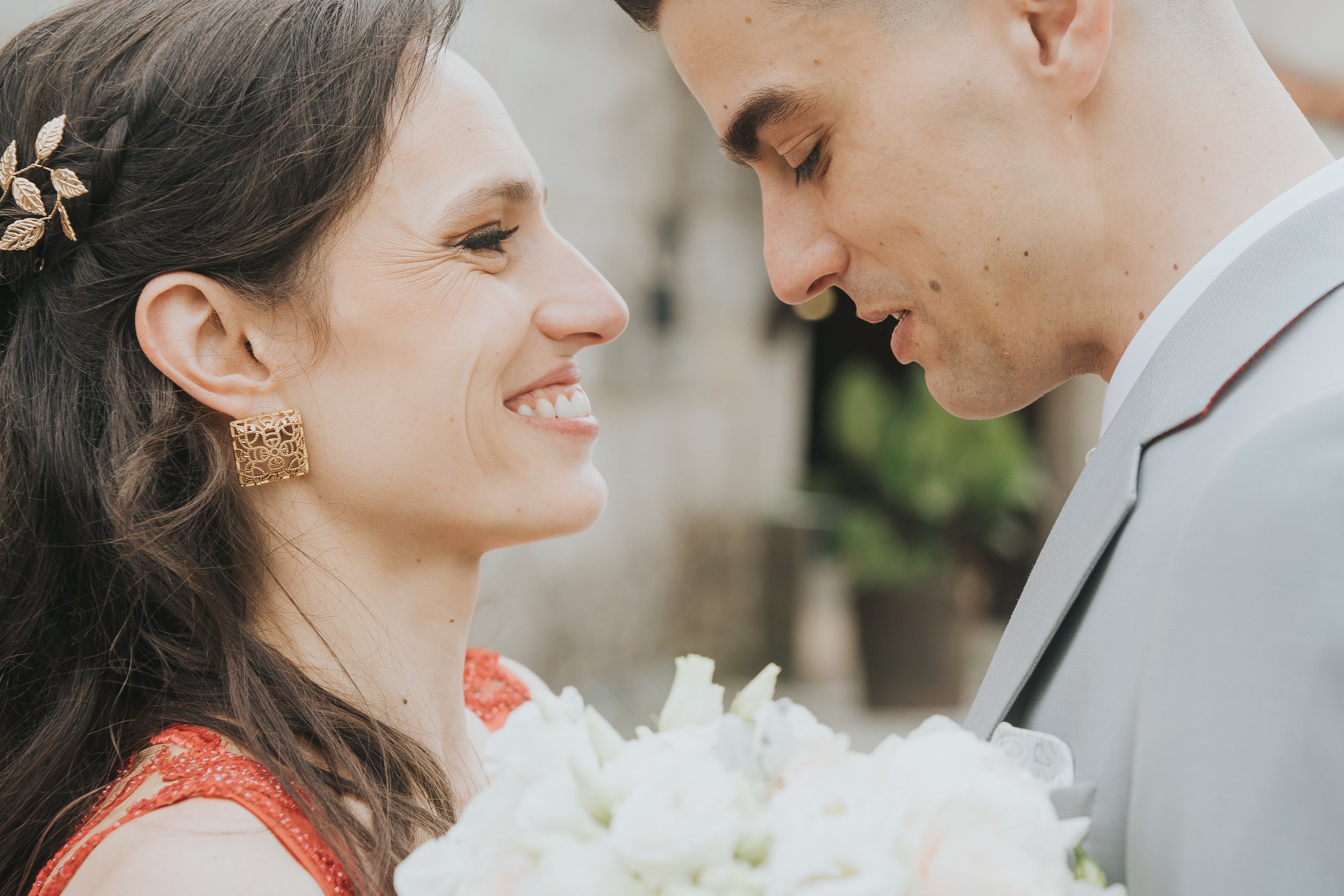 Casal sorrindo frente a frente em ambiente ao ar livre. A mulher usa vestido vermelho e brinco dourado, enquanto o homem veste terno cinza. Ela segura um buquê de flores claras.