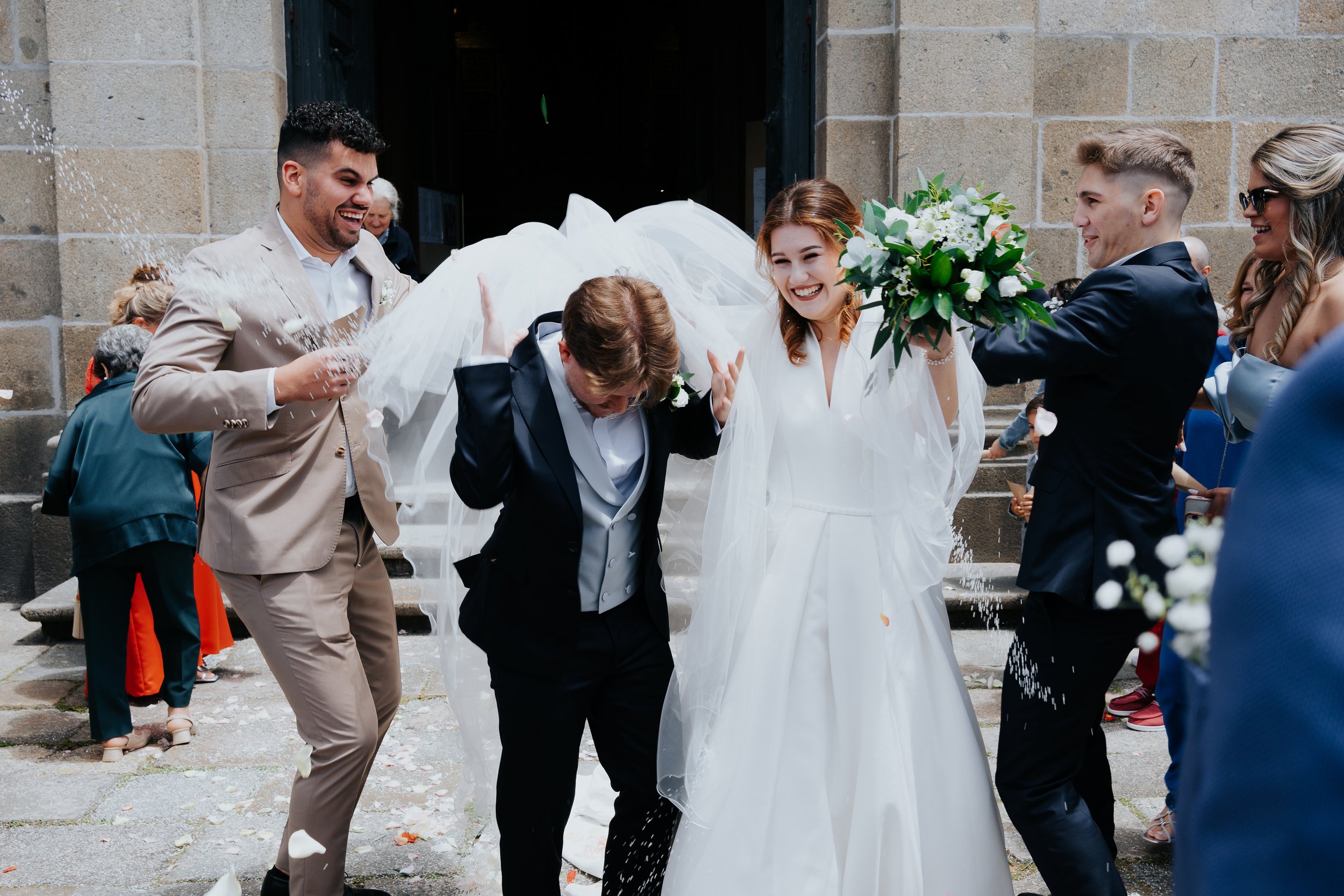 Grupo de pessoas celebrando em um casamento, com noiva sorrindo e segurando um buquê, cercada por convidados jogando pétalas de flores do lado de fora de um edifício.