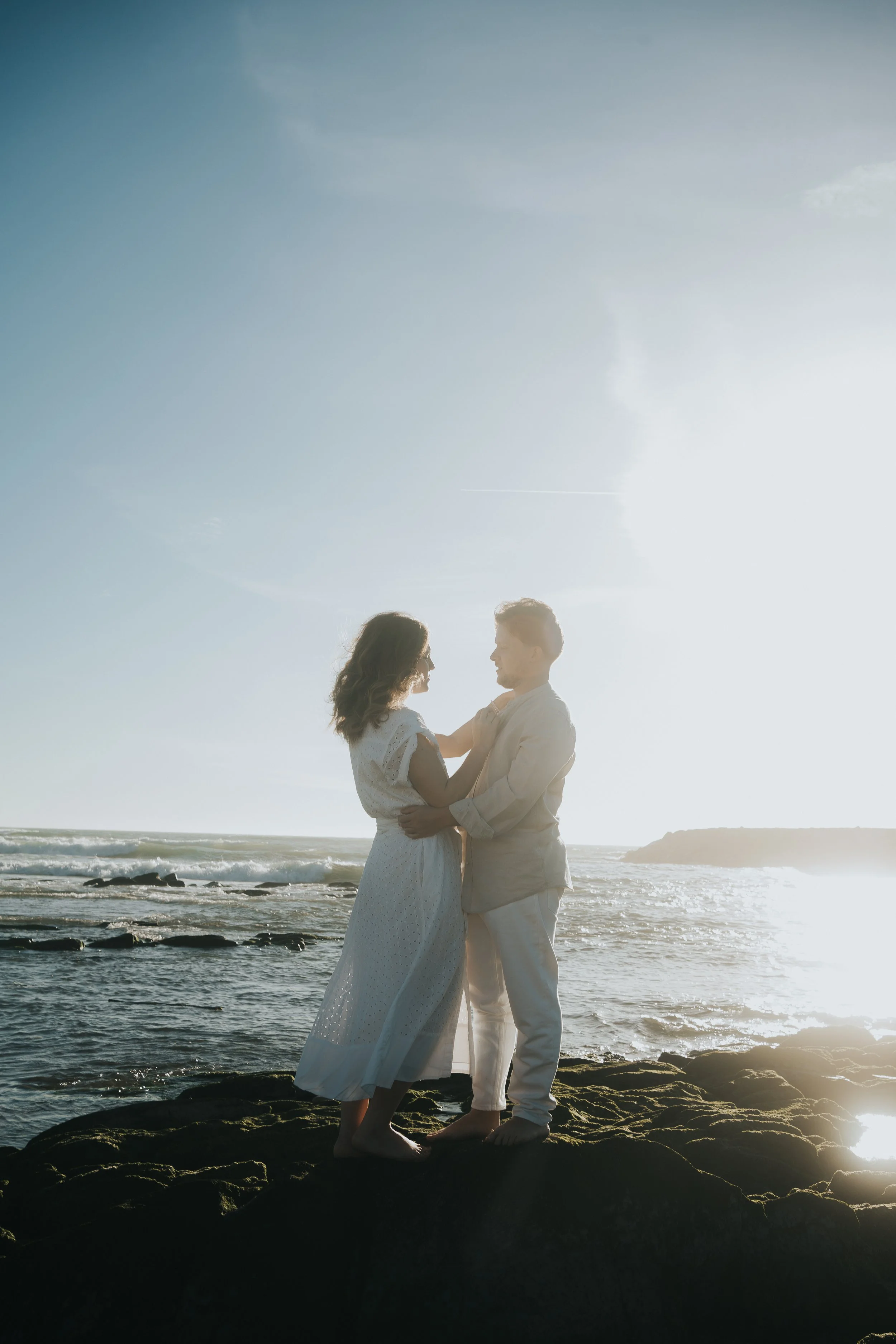 Casal abraçado em pé sobre rochas à beira-mar, com o pôr do sol ao fundo; vestem trajes claros e são iluminados pela luz suave do sol.