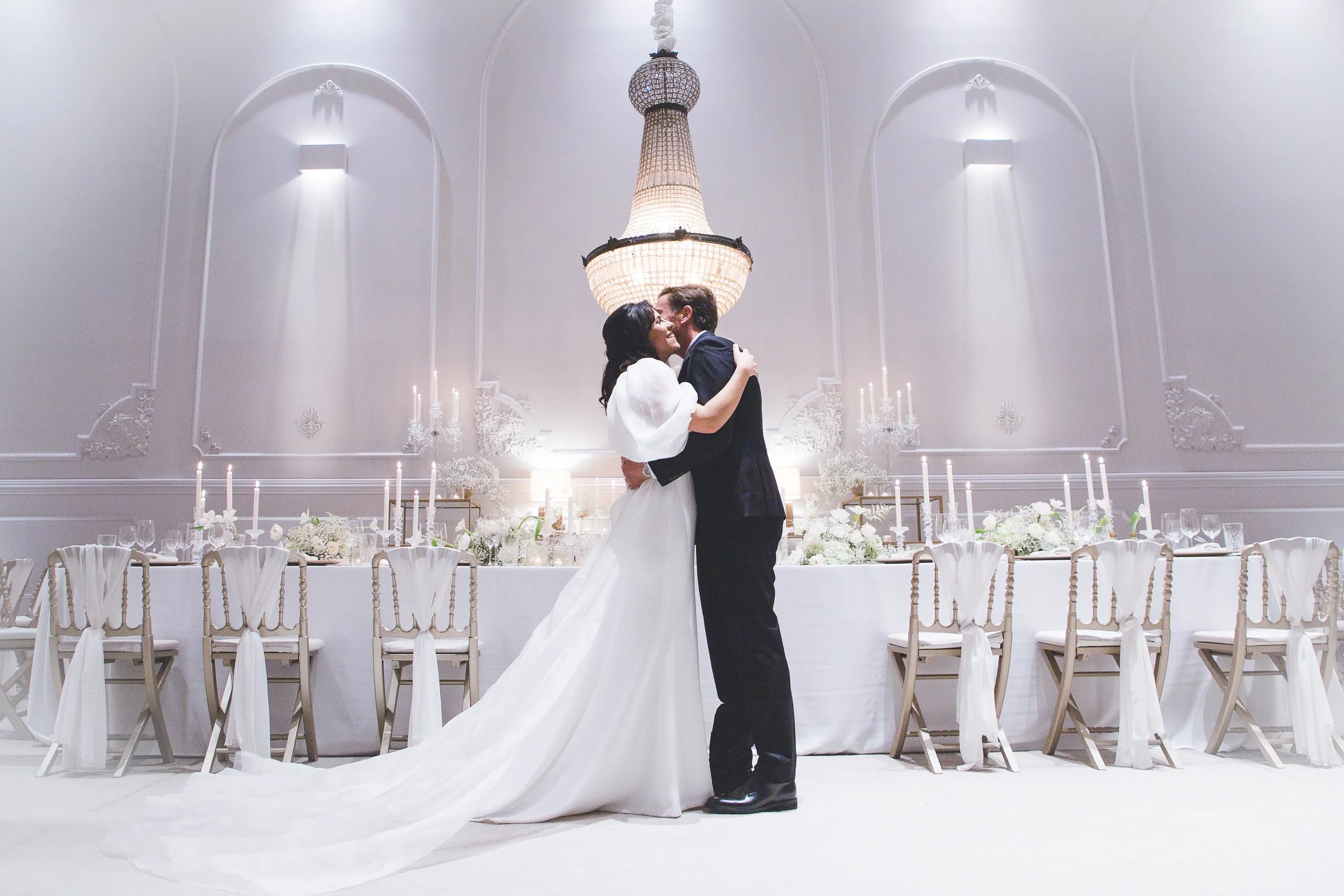 Casal se beijando em um salão de casamento elegante, com decoração branca, mesa arrumada e candelabros.