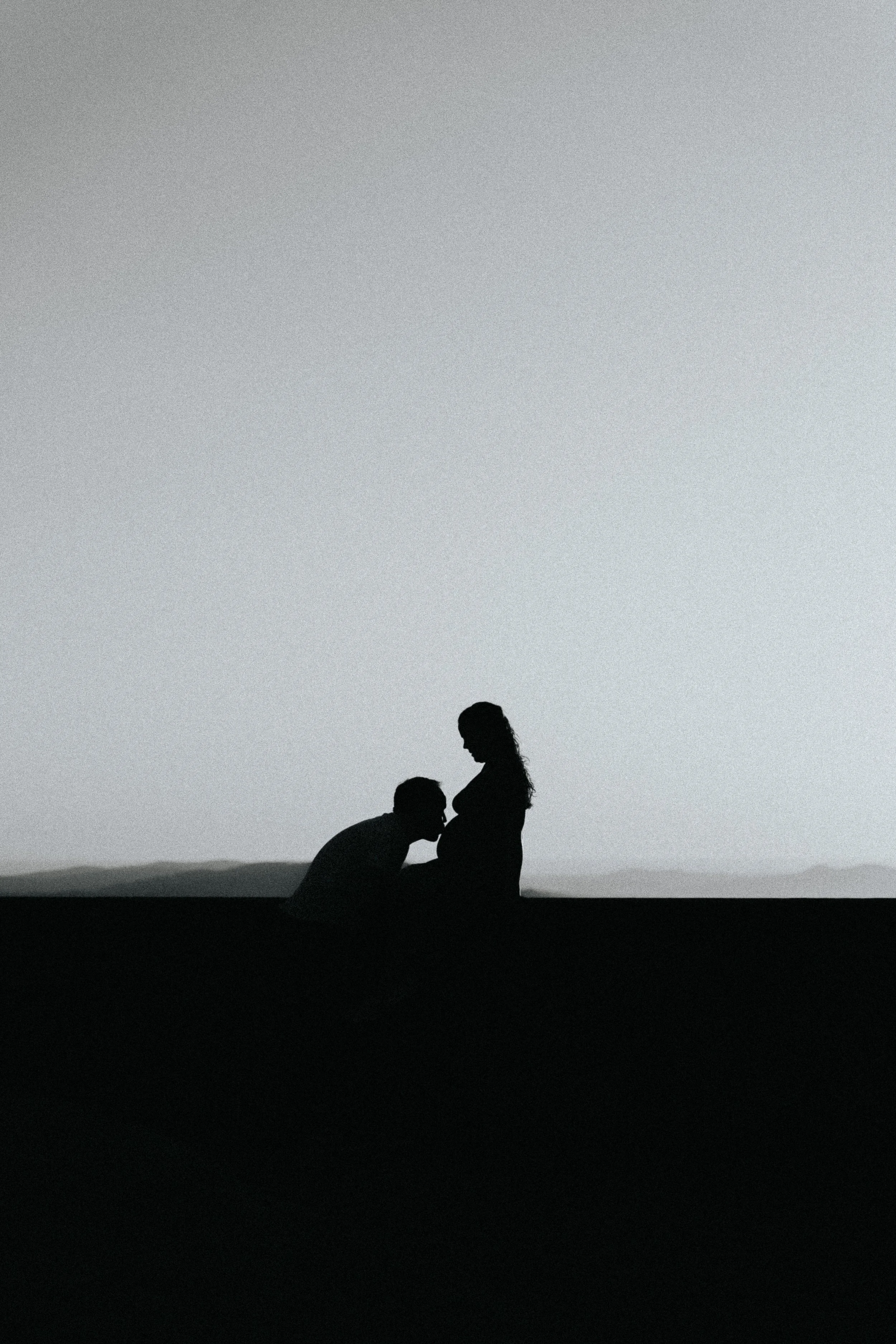 Silhueta de um homem beijando a barriga de uma mulher grávida em um cenário ao ar livre.