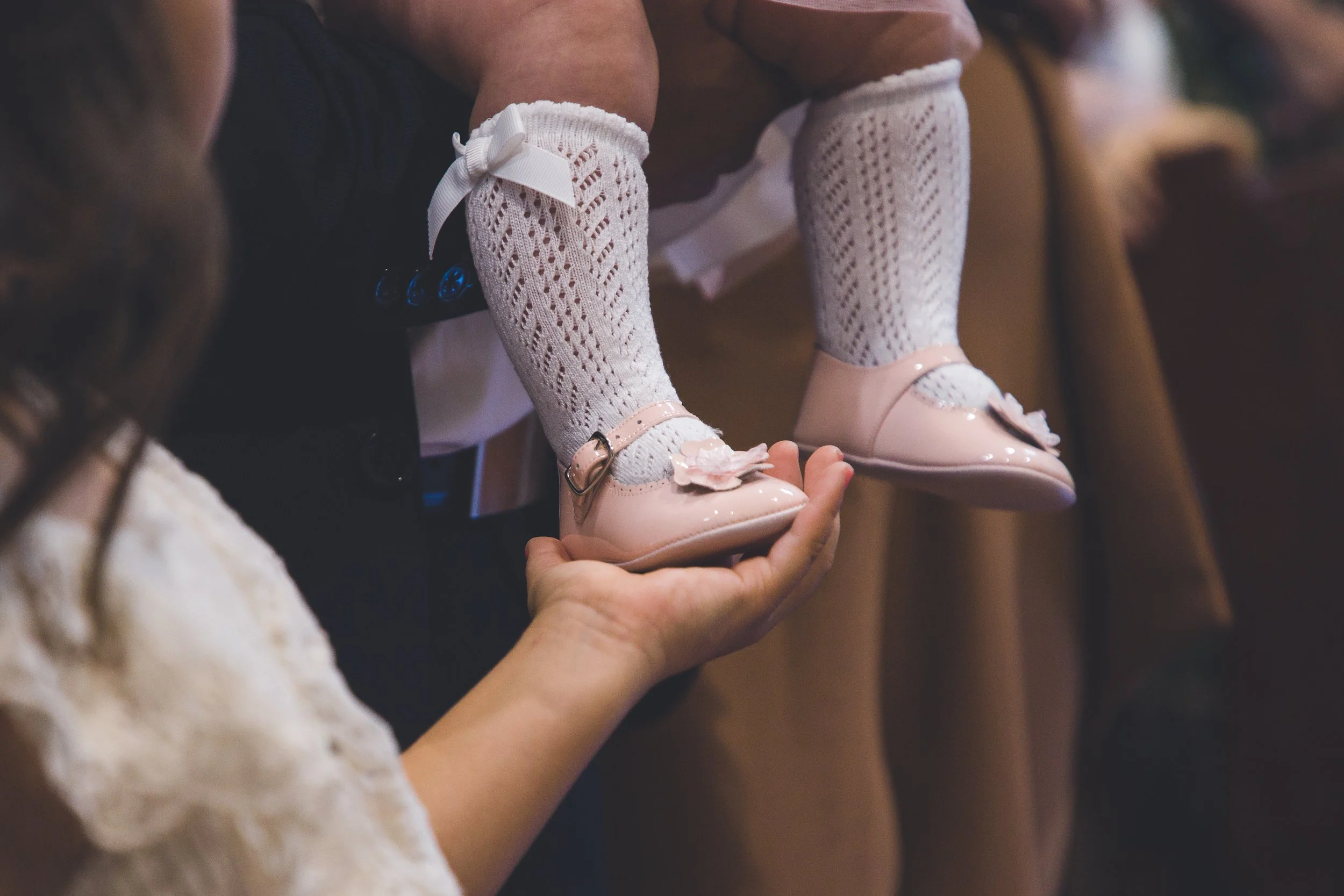 Bebê com meias brancas de renda e sapatos rosa claros decorados com flor, sendo segurado por um adulto.