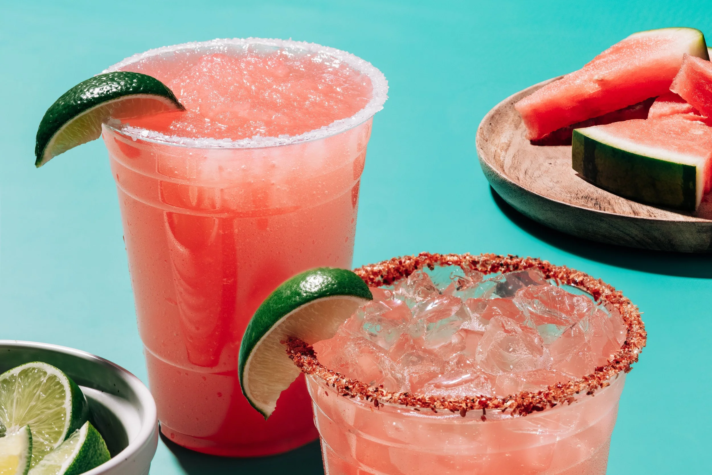 Pink frozen beverage in a glass with a salted rim and lime wedge, garnished with a lime wedge. Surrounding are sliced limes, a bowl of lime wedges, and slices of watermelon on a wooden plate, on a turquoise background.