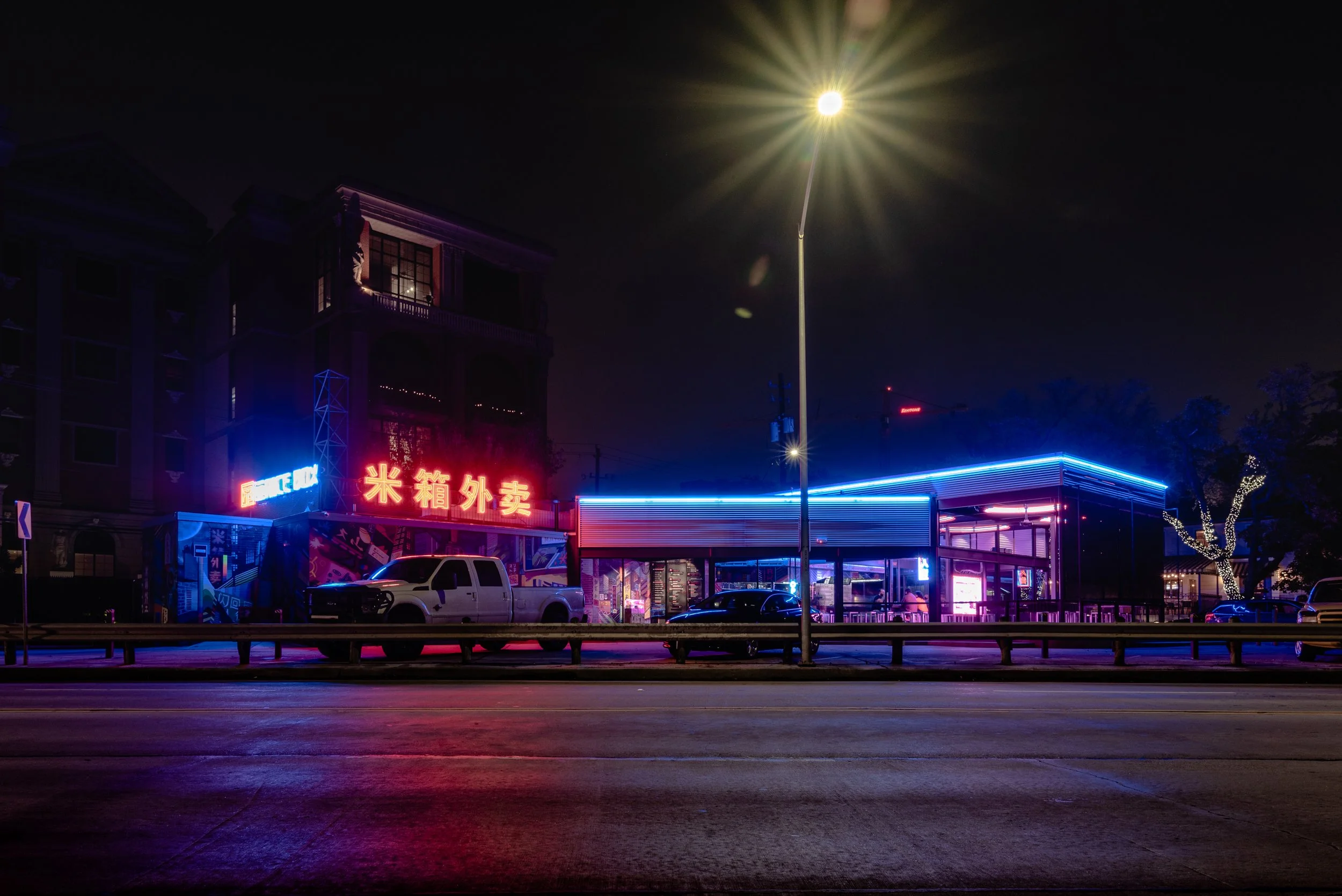Night scene of a commercial area with neon signs, parked cars, and a brightly lit streetlamp.