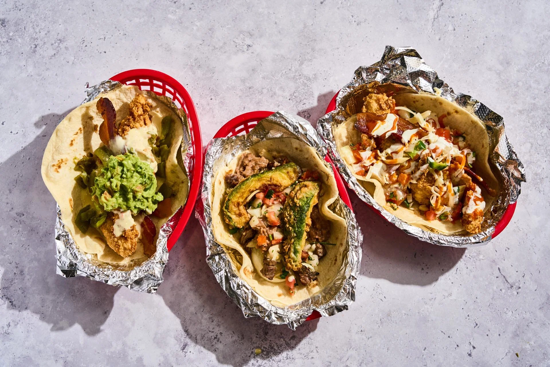 Three tacos in red plastic baskets lined with foil. The tacos contain ingredients such as guacamole, fried chicken, beef, grilled vegetables, sour cream, cheese, and hot sauce, served on soft tortillas. The tacos are placed on a light gray textured s