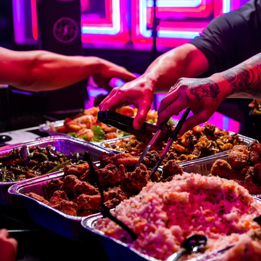 Hands using tongs to serve food from a buffet with bright neon lights in the background.