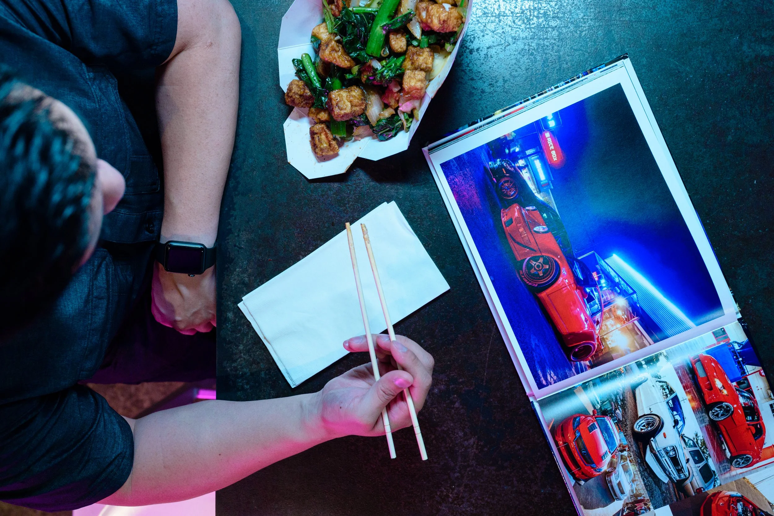 Person sitting at a table with a plate of Asian-style stir-fry containing fried chicken pieces and vegetables, reading a magazine with car images, using chopsticks, wearing a smartwatch, and surrounded by a napkin and chopsticks on a dark textured su