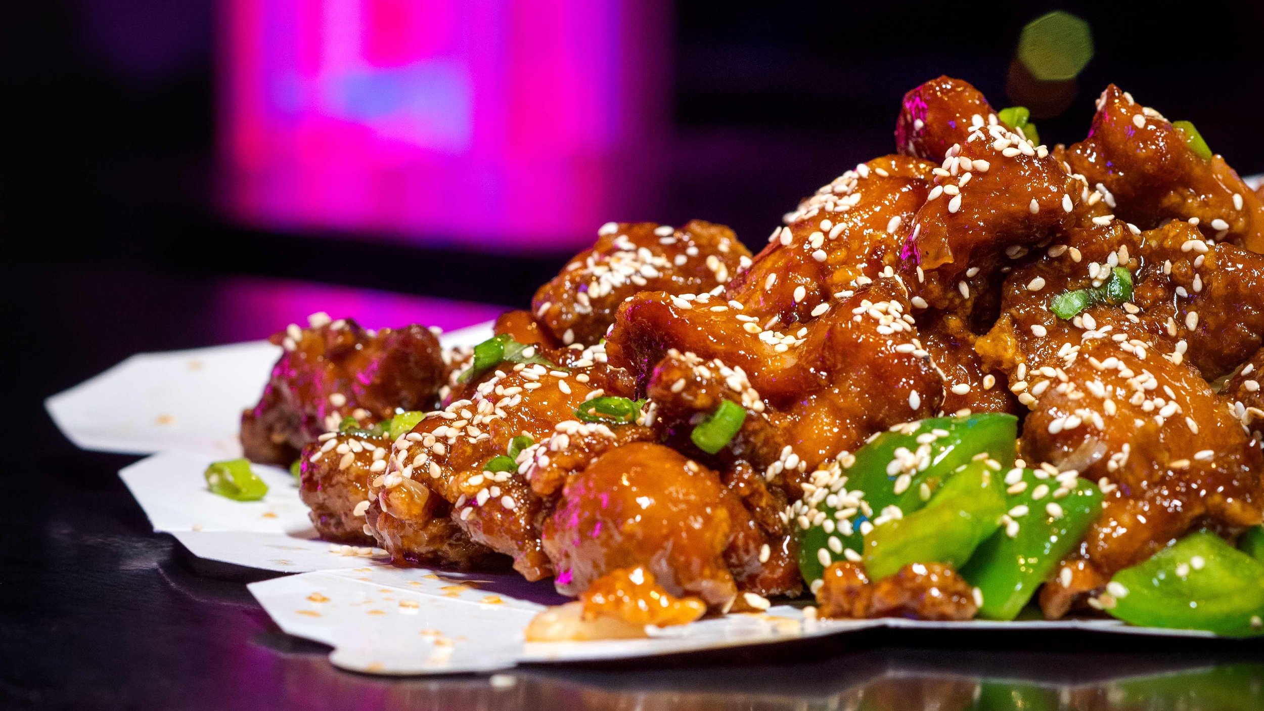 Close-up of glazed chicken pieces garnished with sesame seeds, green onions, and pink edible flowers on a white paper serve tray, with a blurred purple and black background.