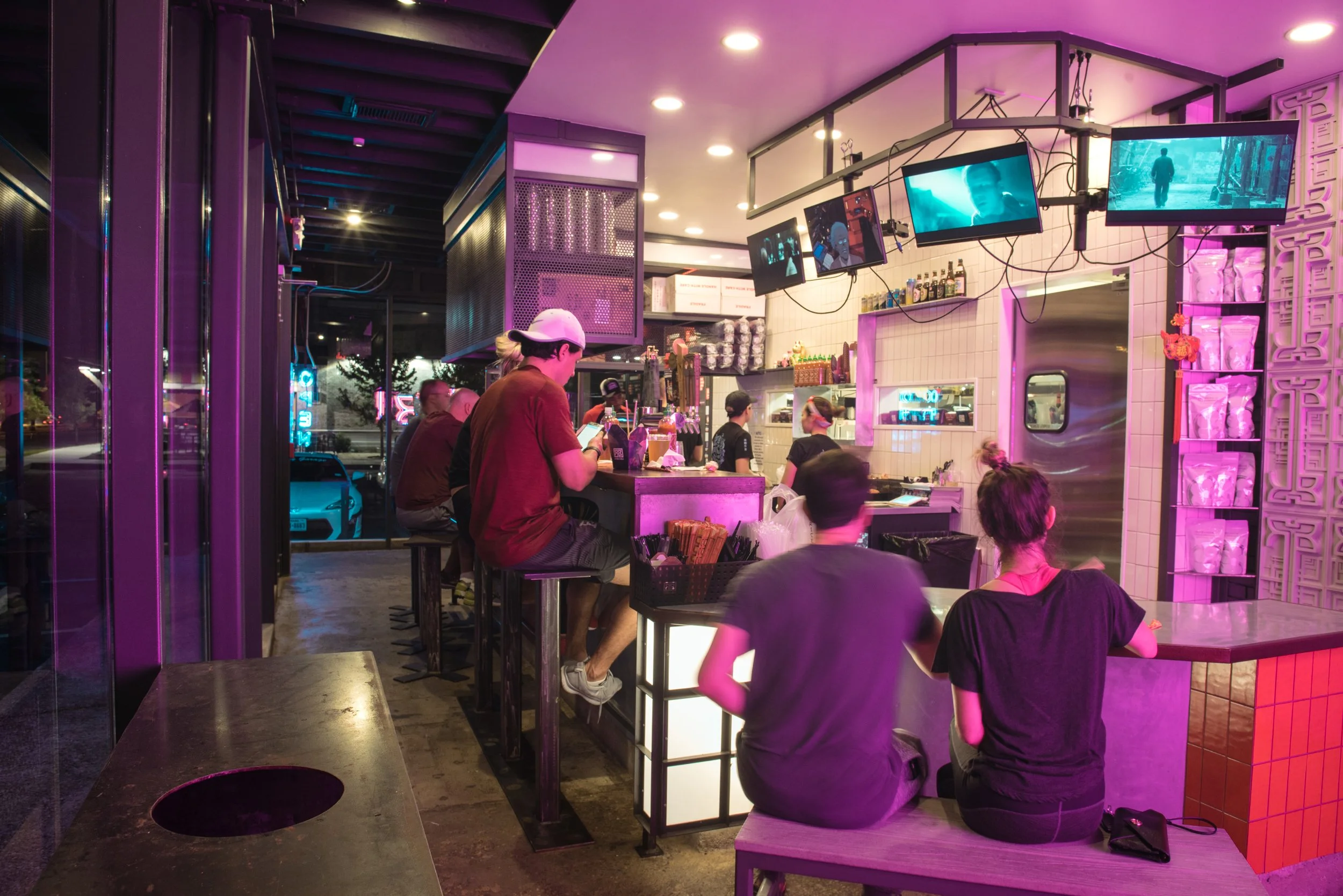 People sitting and waiting inside a modern cafe or bar with purple lighting, multiple TVs on the wall displaying videos, and a counter area with drinks and snacks.
