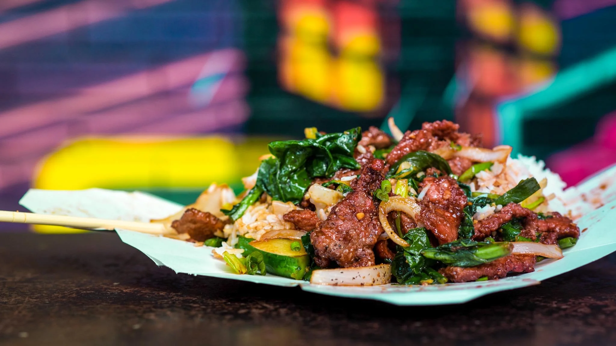 Close-up of a plate of Asian stir-fried beef with vegetables and white rice, served on a green paper tray, with a colorful blurred background.