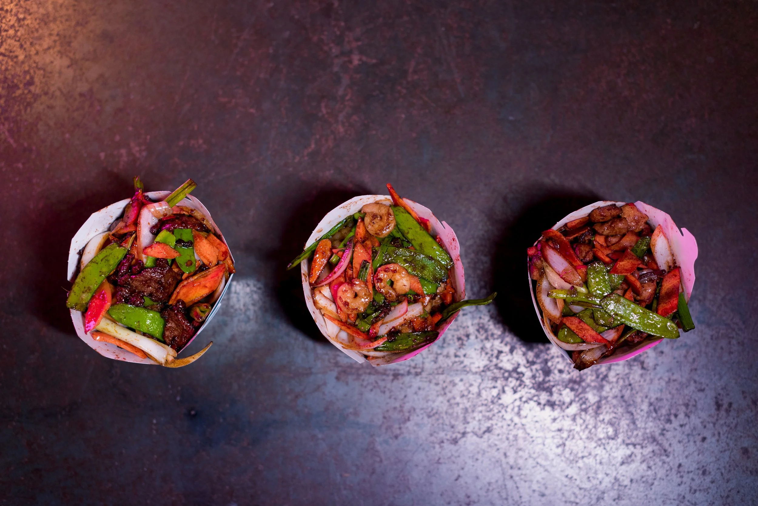Three paper cups filled with mixed vegetables and cooked meat, placed on a dark textured surface.