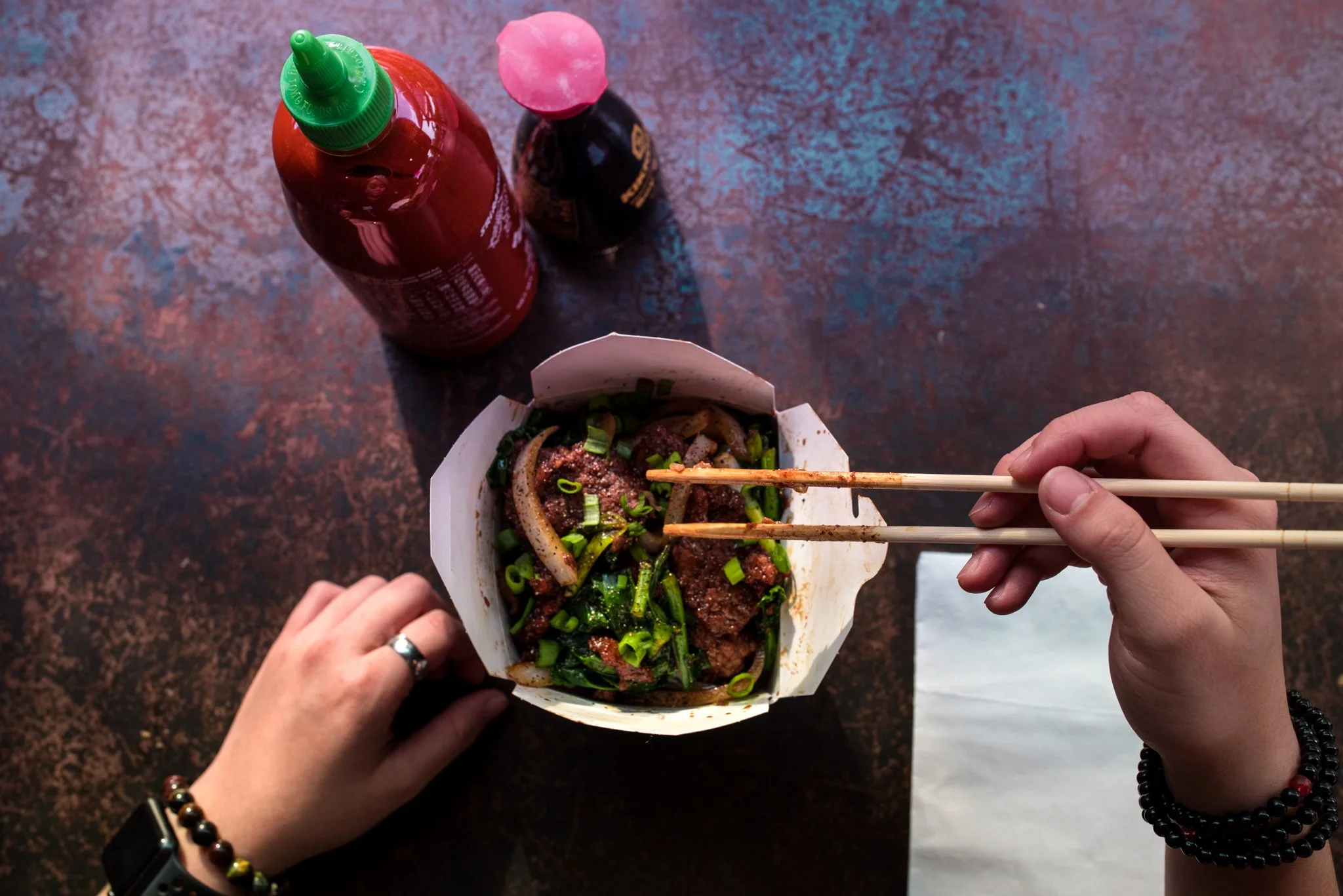 Top-down view of a portable container holding Asian noodles with beef, chopped green onions, and herbs. Two hands are holding chopsticks and a paper container. On a textured surface, there are two bottles, one with a green cap and one with a pink cap