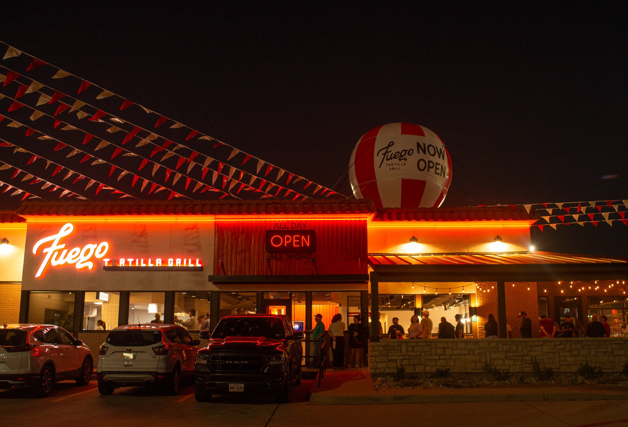Nighttime scene of a taco restaurant with a sign reading 'Fuego' and 'Tortilla Grill', illuminated storefront, parked cars in front, and a hot air balloon with red and white stripes that says 'Fuego NOW OPEN' in the background.