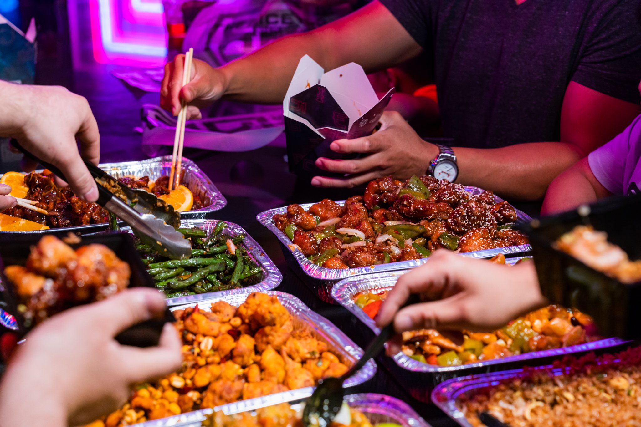 People serving themselves food at a buffet with Asian dishes, including vegetables and meat, in disposable aluminum trays.