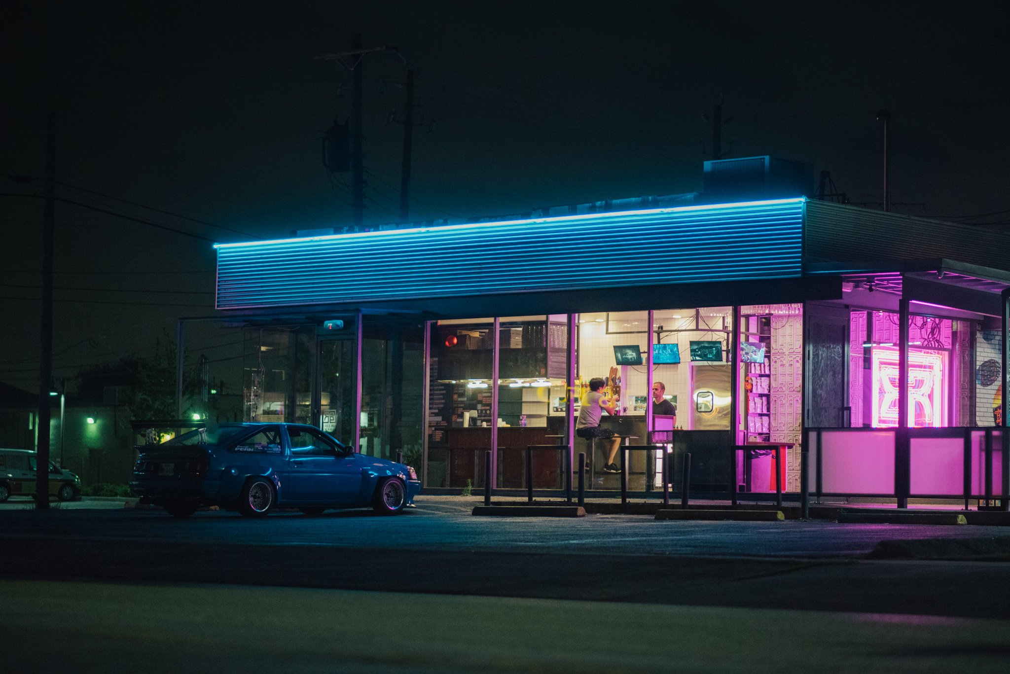 Nighttime scene of a brightly lit neon diner with pink and blue signage, visible through large windows, with two people sitting at a counter inside and a parked blue car outside.