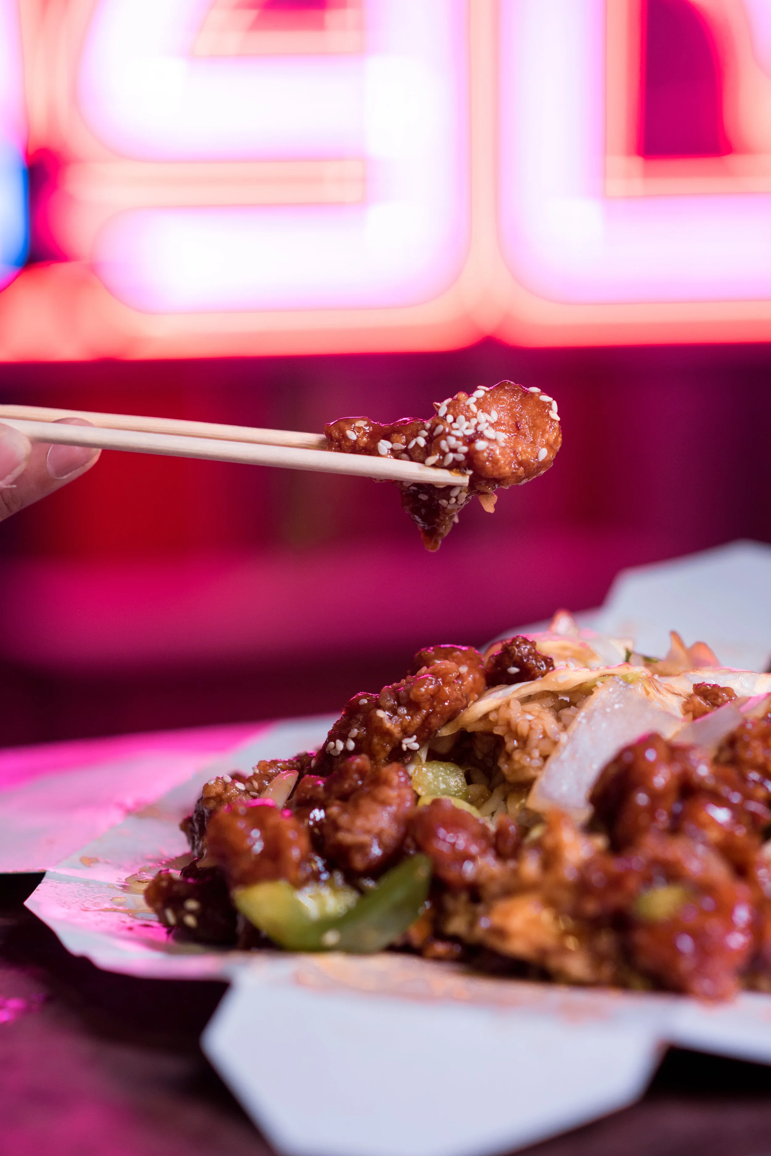 Close-up of a piece of sushi with fried chicken, sesame seeds, and vegetables on a plate, with vibrant pink neon lighting in the background.