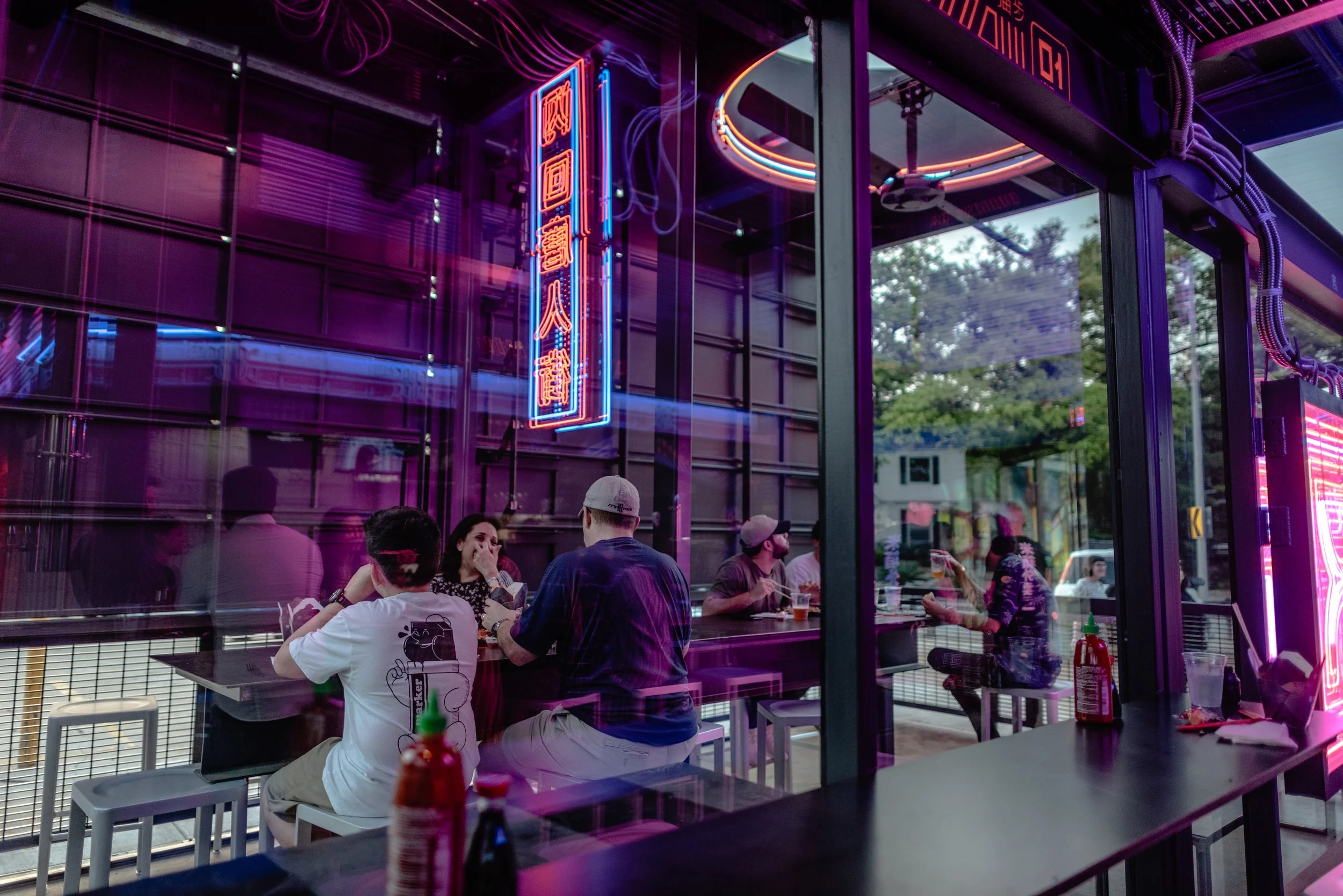 A group of people sitting at tables inside a neon-lit restaurant, with purple and pink neon signs and decorations, and some trees visible outside through the glass window.