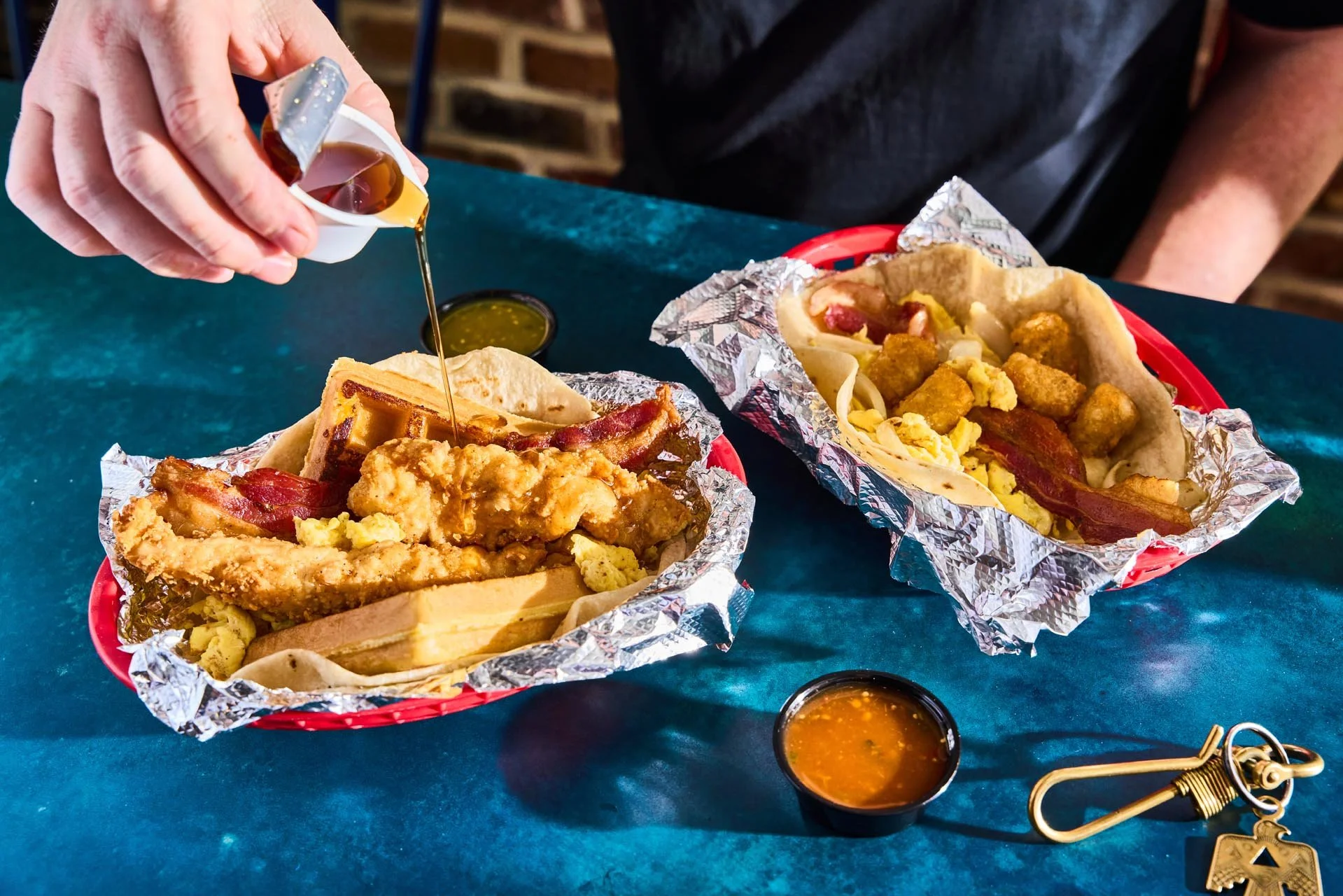 Two foil-lined baskets on a teal table, each containing a breakfast sandwich with fried chicken, bacon, and scrambled eggs, with small containers of syrup and hot sauce nearby.