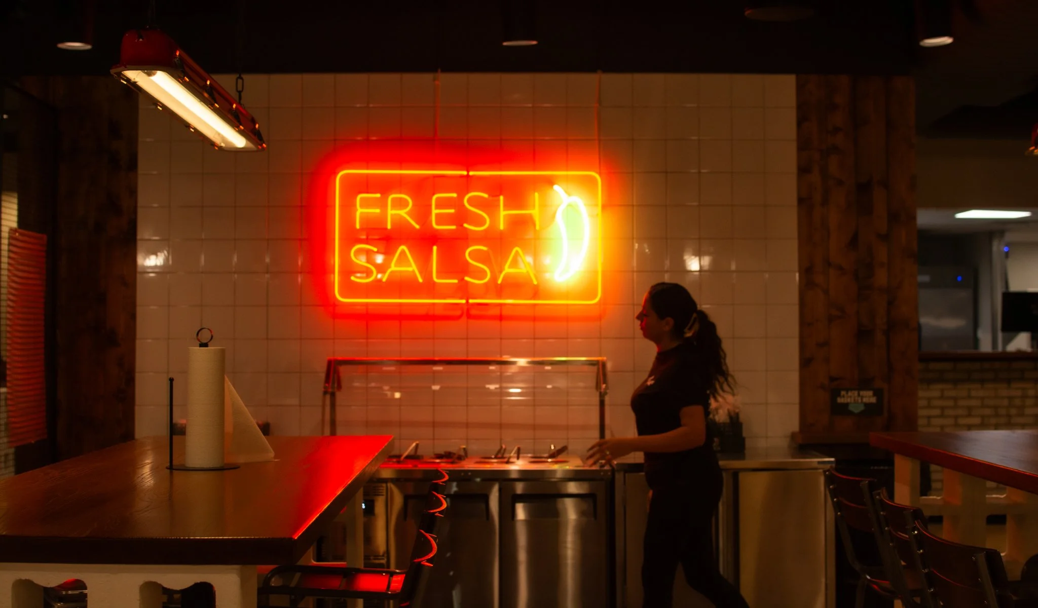 Neon sign reading 'FRESH SALSA' with a chili pepper graphic, inside a restaurant with a woman standing at a counter and tables in the foreground.