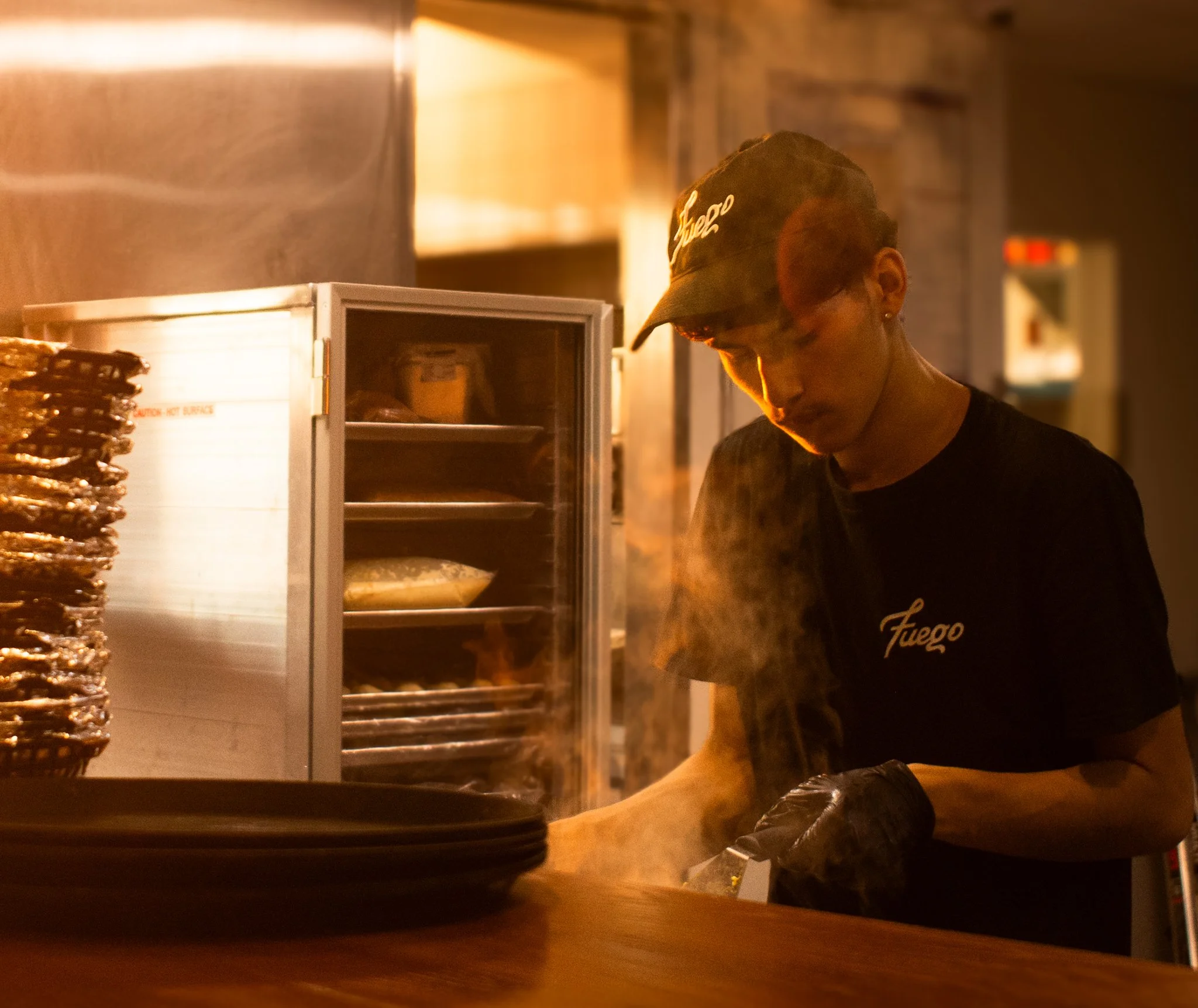 A person wearing a black cap with white writing and glasses working behind a counter with a small refrigerator, a stack of plates, and a plate with grilled food at a restaurant.