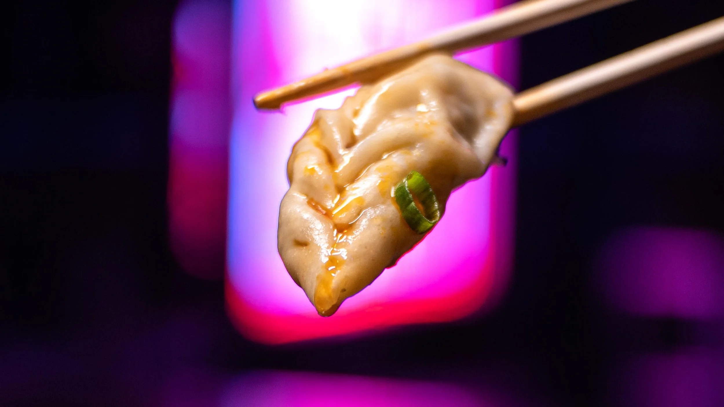 A close-up photograph of a slice of pizza being held by chopsticks against a colorful purple and pink background.