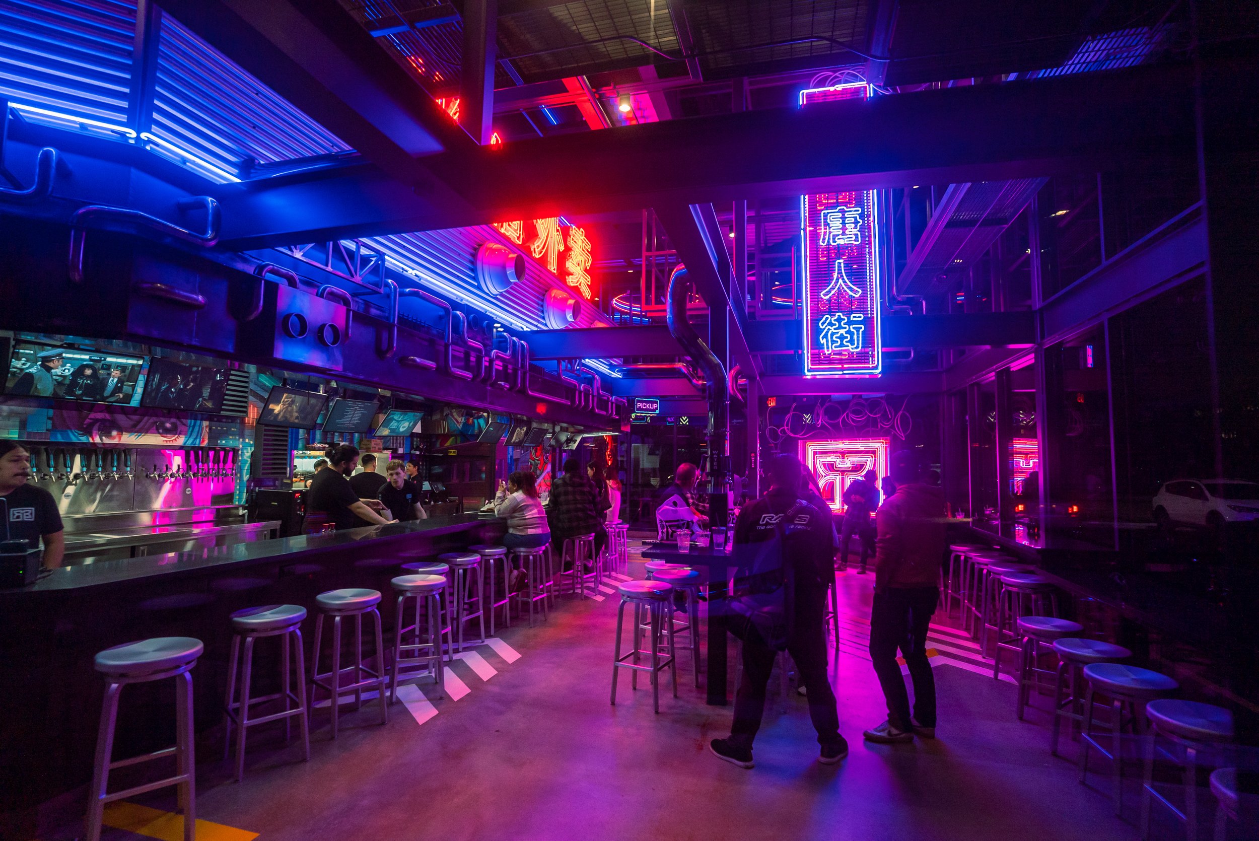 Interior of a vibrant bar or restaurant with neon pink and blue lights and several customers seated at the bar and high tables.