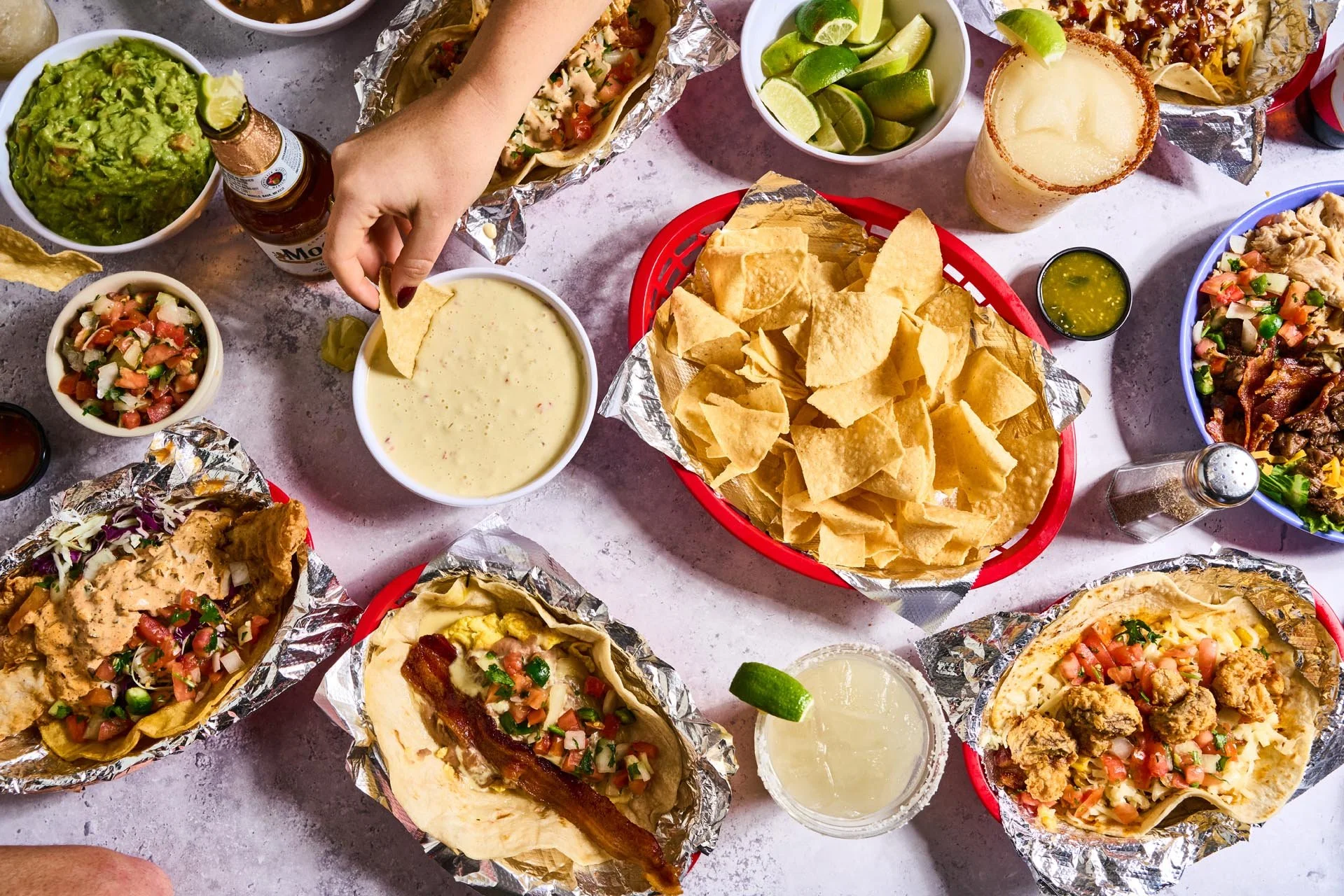 An overhead view of a Mexican meal spread on a table with tacos, nachos, guacamole, salsa, lime wedges, and drinks, with one hand reaching to pick up a chip.