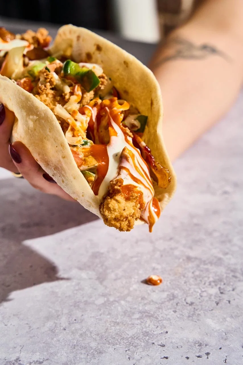 Close-up of a person holding a taco filled with fried chicken, lettuce, shredded cheese, and sauces, with some sauce spilling onto a gray surface.