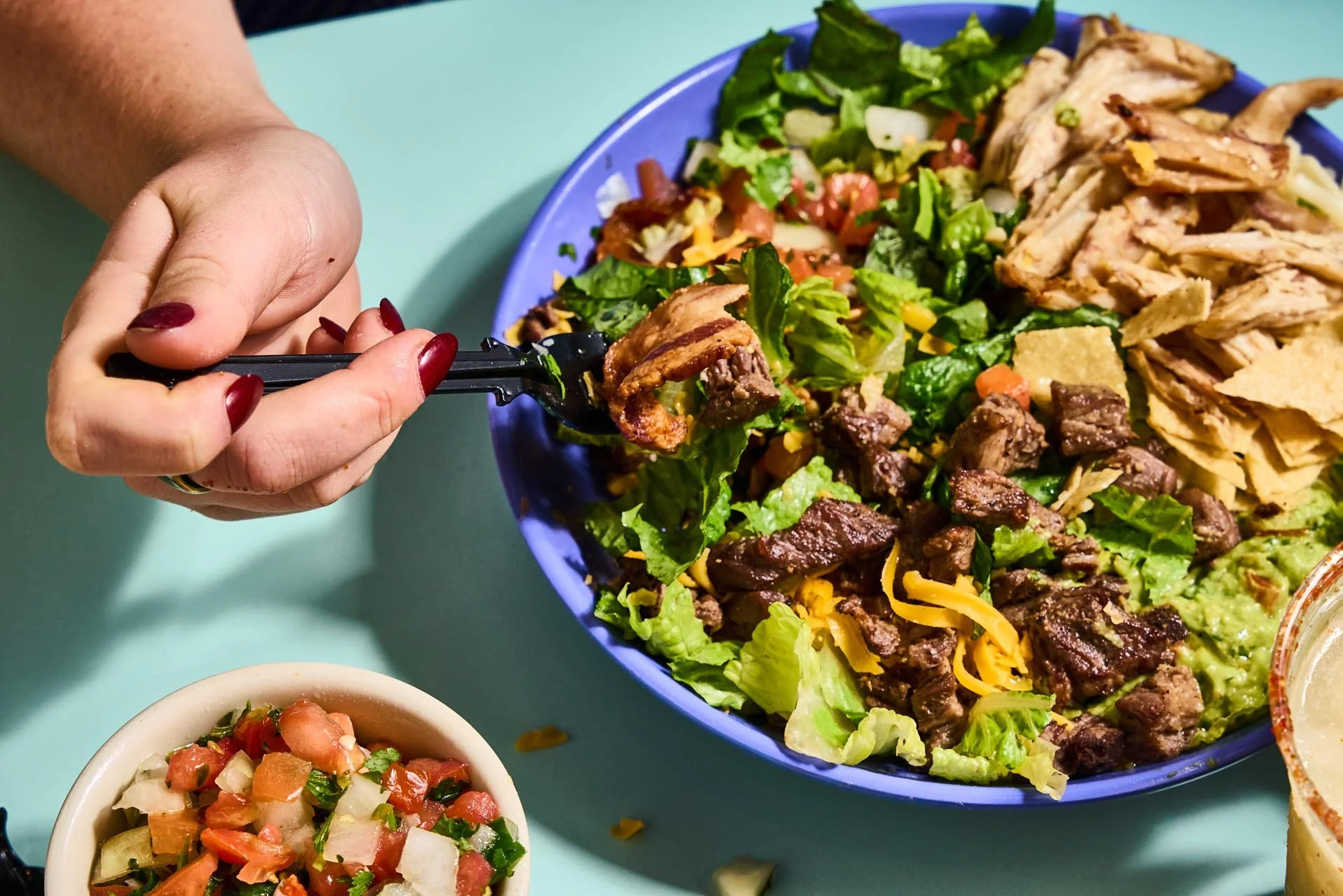 Person with painted nails scooping a salad with lettuce, tomatoes, and shredded cheese from a blue bowl. A smaller bowl of tomato and cucumber salad is visible in the foreground, and part of a yellow dish is visible on the right.