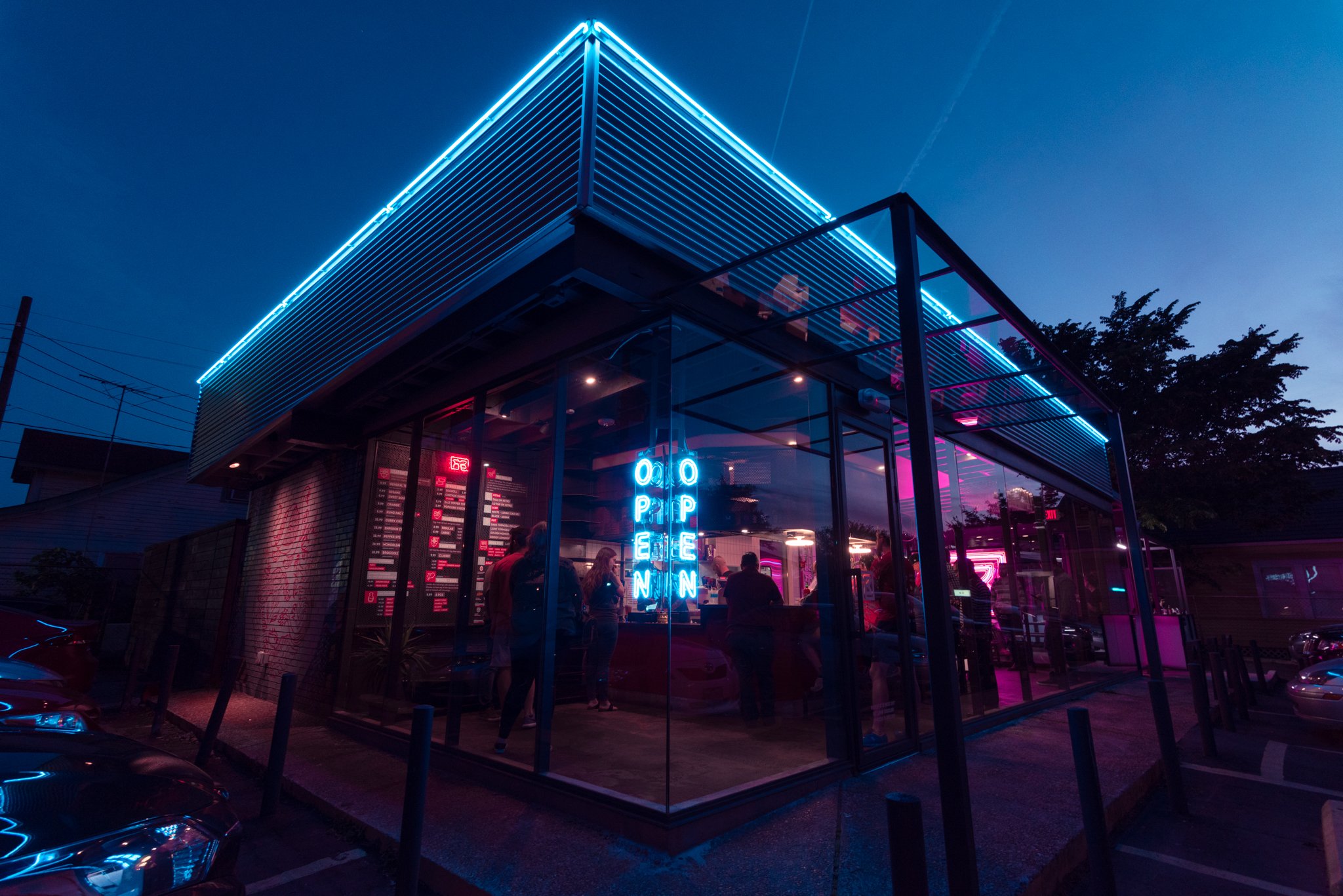 Modern restaurant with blue and pink neon lights, glass exterior, and people inside, visible through the glass walls, during evening or night time.