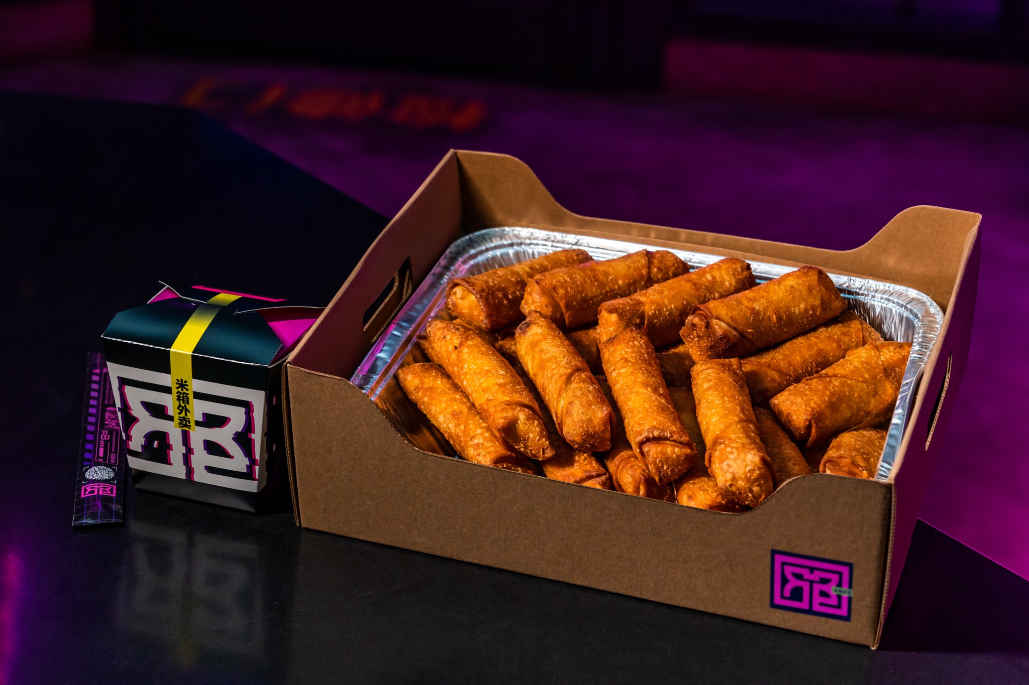 A black takeout box with a tray of fried spring rolls inside. A container with Asian characters and a black packet are positioned next to the box, set on a dark surface with purple lighting.