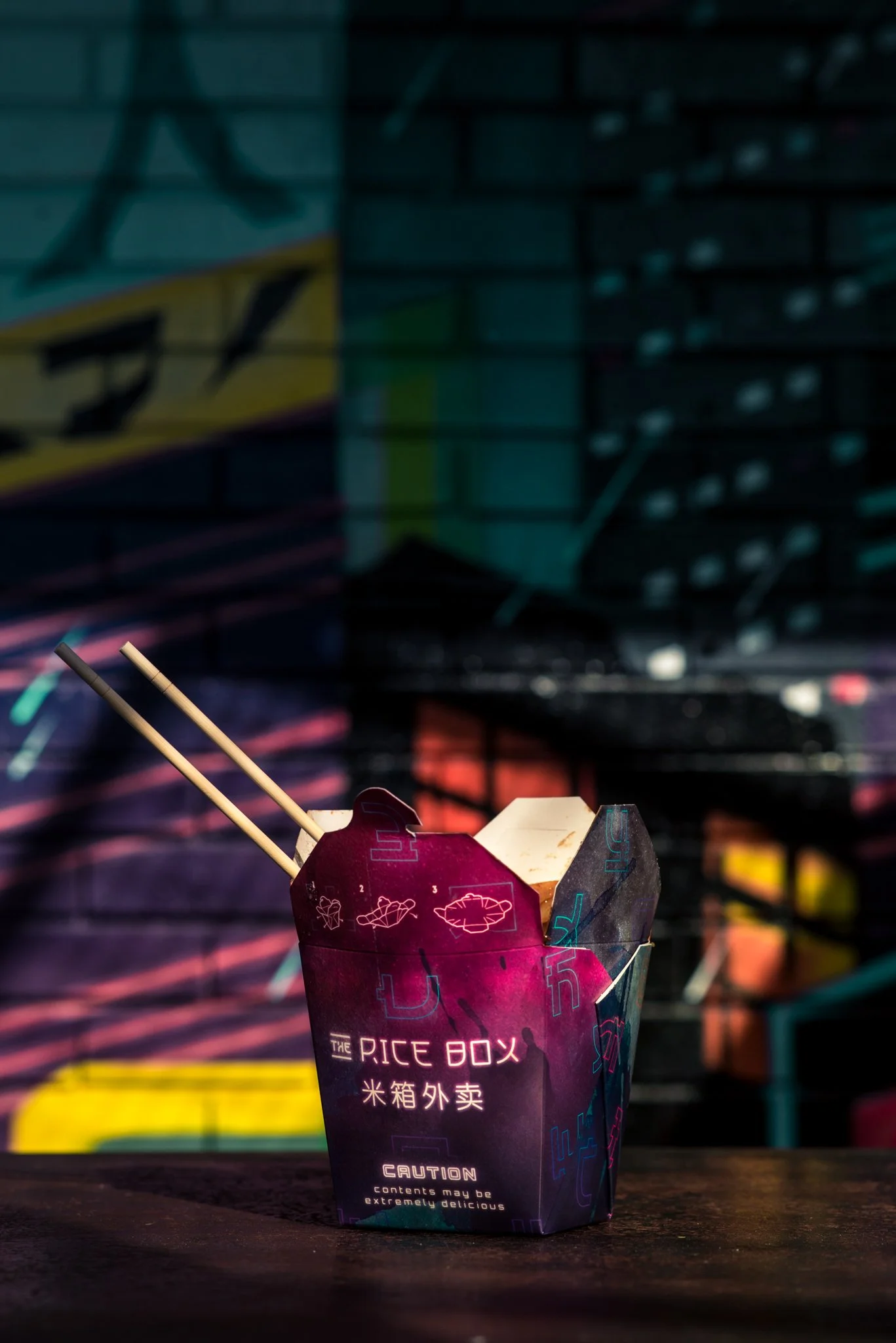 A takeout box labeled "The Rice Box" with chopsticks on top, set against a colorful, blurred cityscape background with neon lights.