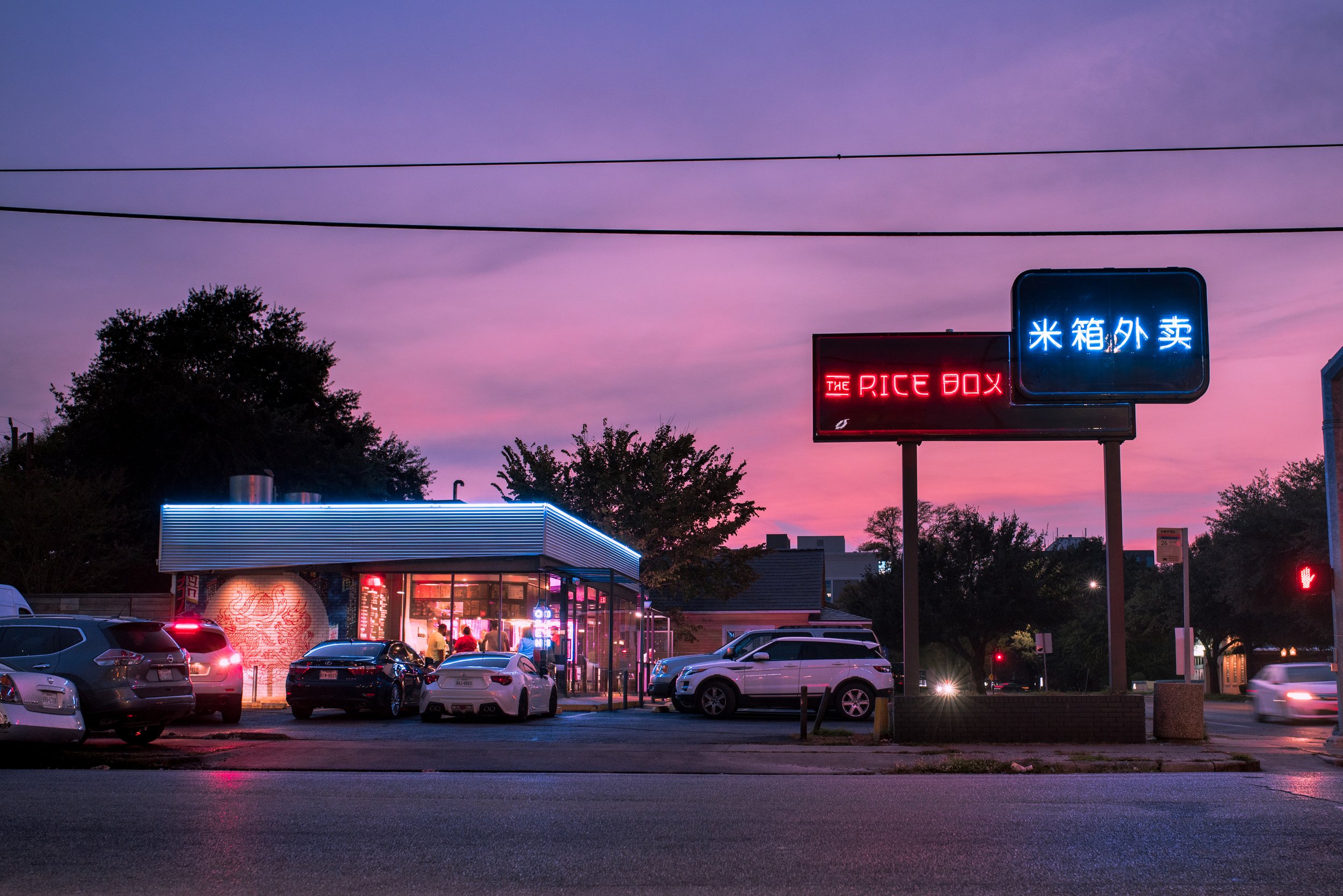 Neon signs outside a small restaurant called 'The Rice Box' against a purple sunset sky, with parked cars and people inside visible through windows.