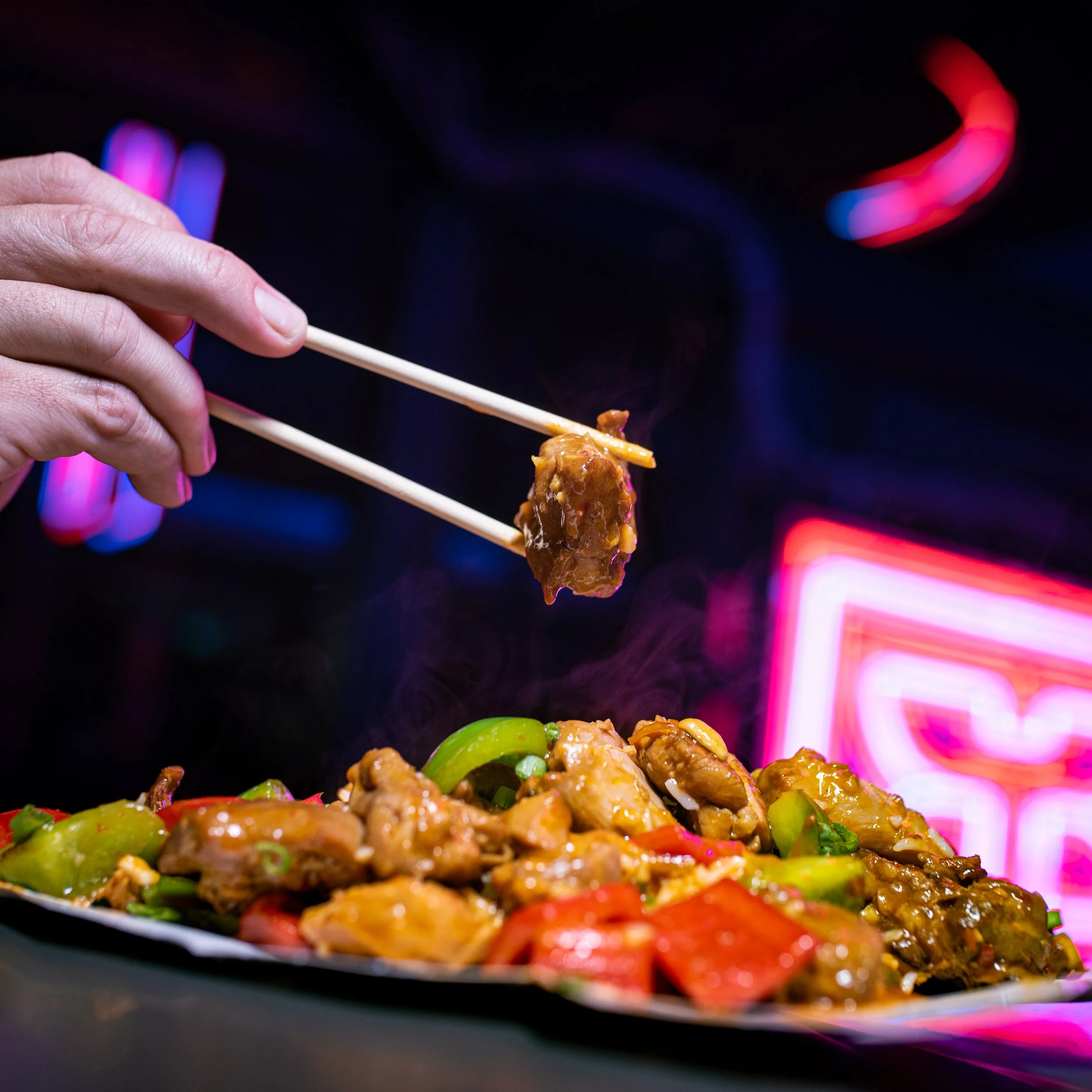 Close-up of a hand holding chopsticks with food, with a blurred background of neon lights in red and blue.