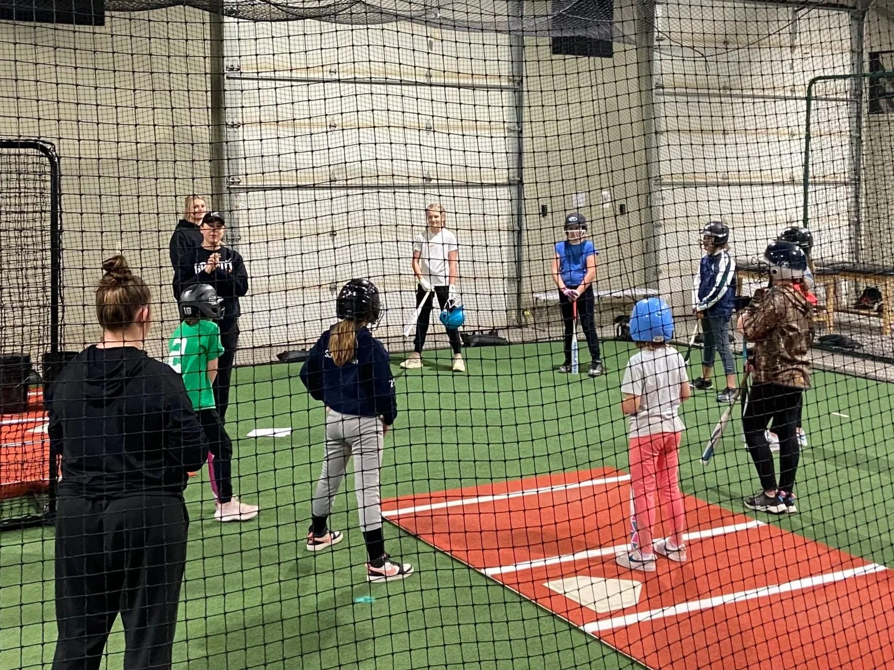 Children participating in a softball camp inside a batting cage. Many children wearing helmets, with a trainer and instructors guiding them.
