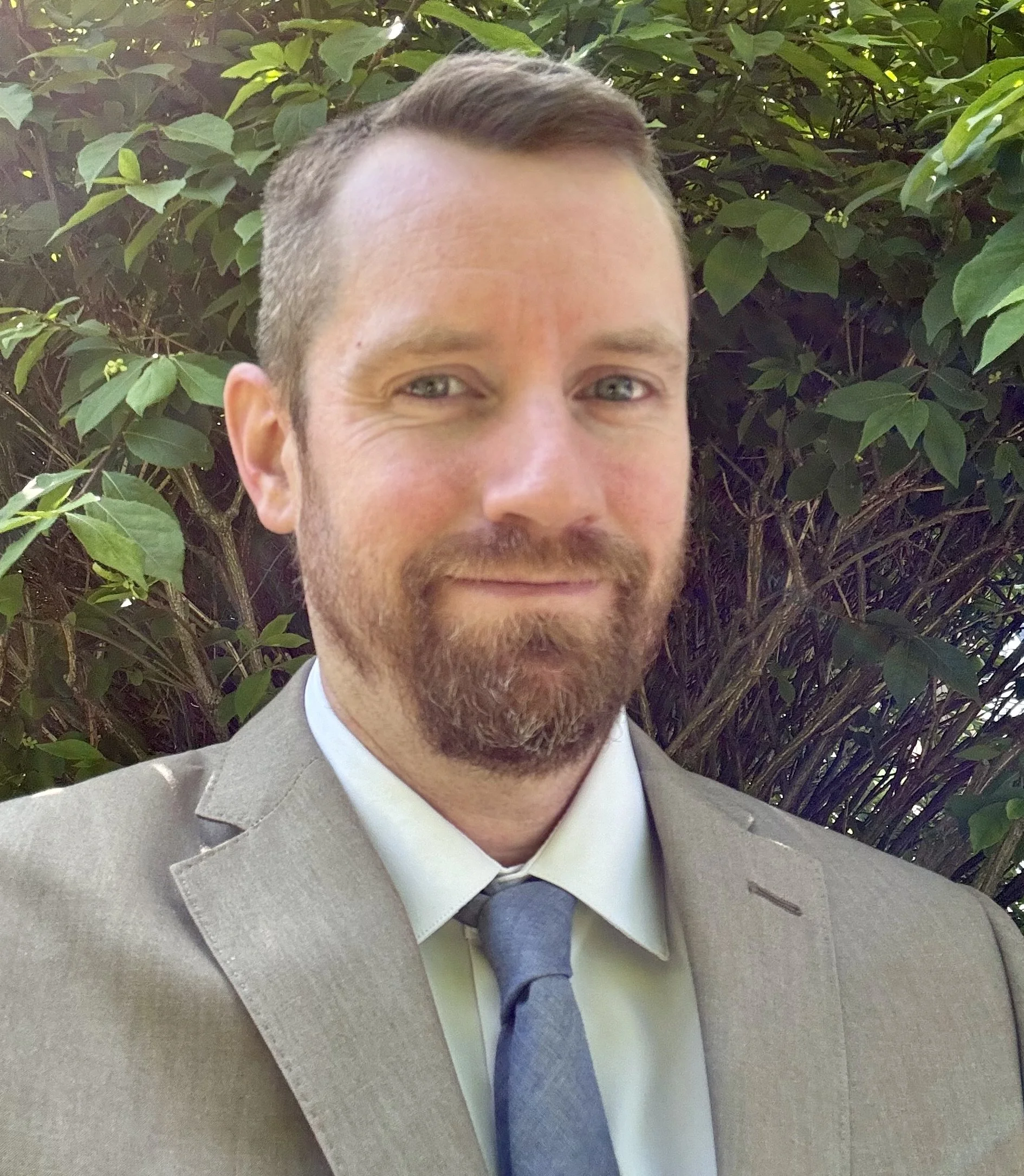 Sean Diamond in a tan suit and light blue necktie standing in front of a bush with green leaves.